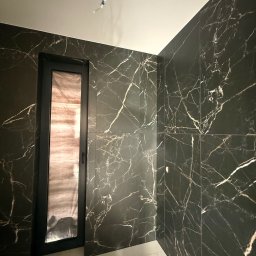 KARLITO WYKOŃCZENIA WNĘTRZ ADRIAN GŁUSZCZAK - Elegancka łazienka z czarnymi płytkami imitującymi marmur z białymi żyłami, czarna rama drzwi, biało-złota podłoga. Widoczne niedokończone podłączenie oświetlenia.