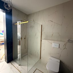 KARLITO WYKOŃCZENIA WNĘTRZ ADRIAN GŁUSZCZAK - Wykończona łazienka z kabiną prysznicową typu walk-in, białą toaletą i płytkami imitującymi marmur z złotymi akcentami. Widoczne niedokończone oświetlenie.