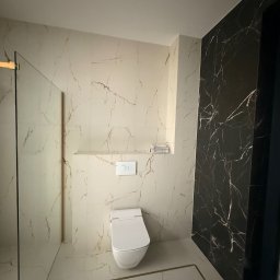 KARLITO WYKOŃCZENIA WNĘTRZ ADRIAN GŁUSZCZAK - Elegancka łazienka z marmurowymi płytkami w odcieniach bieli i czerni, minimalistyczna toaleta, szklana kabina prysznicowa z złotymi akcentami i wnęka z półką.