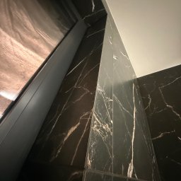 KARLITO WYKOŃCZENIA WNĘTRZ ADRIAN GŁUSZCZAK - Ciemna, marmurowa ściana z widocznym fragmentem okna i sufitu. Ułożenie płytek tworzy geometryczny wzór. Wykończenie wnętrza w nowoczesnym stylu.