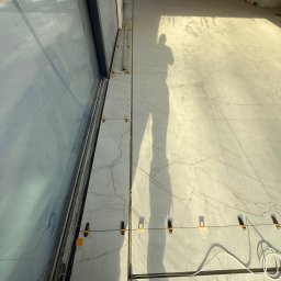 KARLITO WYKOŃCZENIA WNĘTRZ ADRIAN GŁUSZCZAK - Układanie płytek podłogowych z użyciem systemu poziomowania. Widoczny cień osoby, duże okno i narzędzia glazurnicze. Jasne płytki z marmurkowym wzorem.