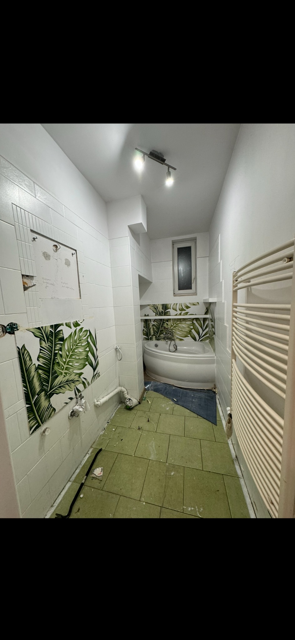 Łazienka w trakcie demontażu: częściowo usunięte białe płytki z motywem liści, odsłonięte rury, zielona podłoga z pozostałościami gruzu, wanna narożna z zabezpieczeniem, grzejnik drabinkowy...