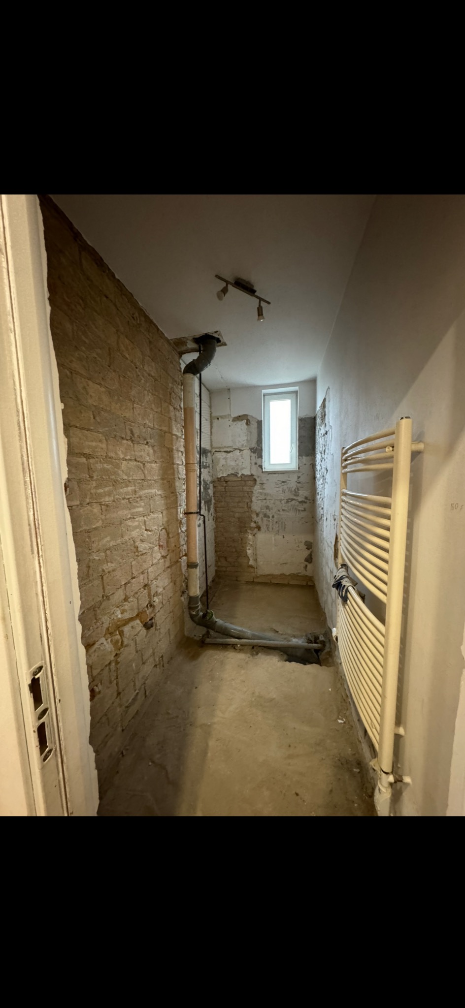 Łazienka w trakcie demontażu: odsłonięta cegła, rury instalacyjne, grzejnik drabinkowy, resztki tynku na ścianach, okno, surowe podłoże.