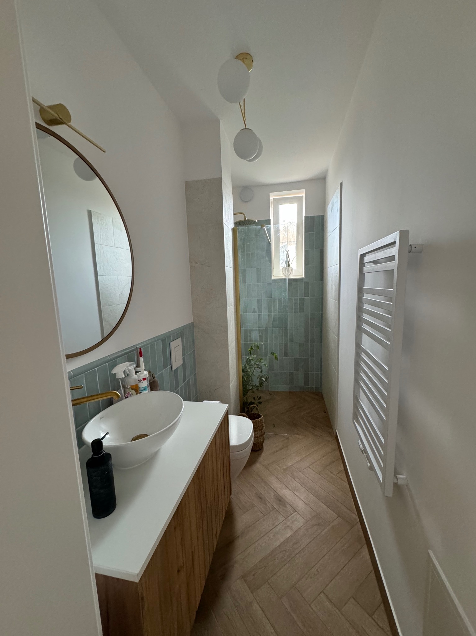 Nowoczesna łazienka z umywalką nablatową, drewnianą szafką, okrągłym lustrem z podświetleniem, grzejnikiem drabinkowym i kabiną prysznicową z zieloną glazurą oraz podłogą ułożoną w jodełkę.