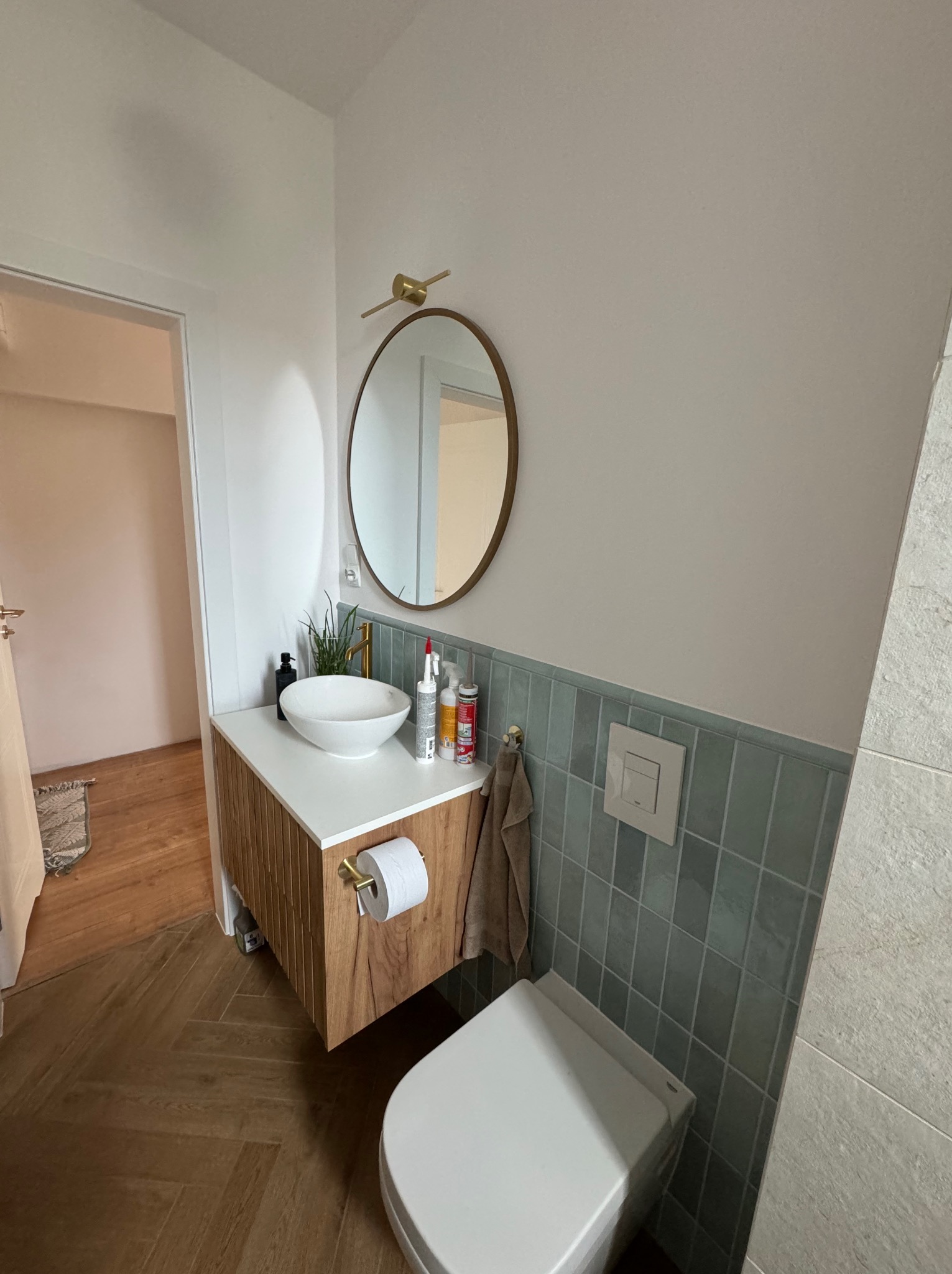 Nowoczesna łazienka po remoncie: umywalka nablatowa na szafce z drewnianymi lamelami, okrągłe lustro z złotą ramą, turkusowe płytki na ścianie do połowy wysokości, podłoga z paneli ułożonych...