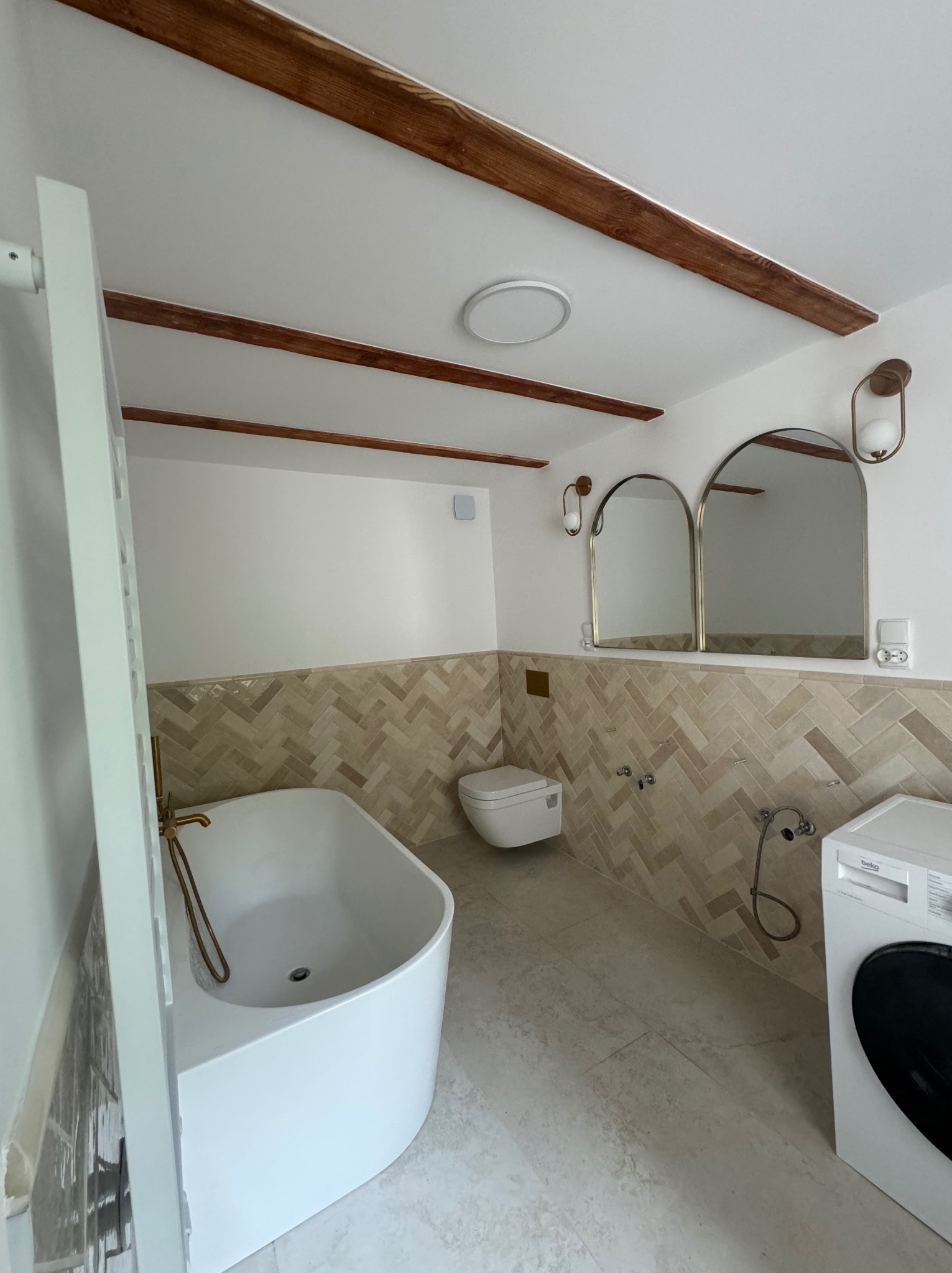 Nowoczesna łazienka z drewnianymi belkami stropowymi, białą wolnostojącą wanną z złotą armaturą, dwoma lustrami w kształcie łuku i płytkami ułożonymi w jodełkę, pralka Beko.