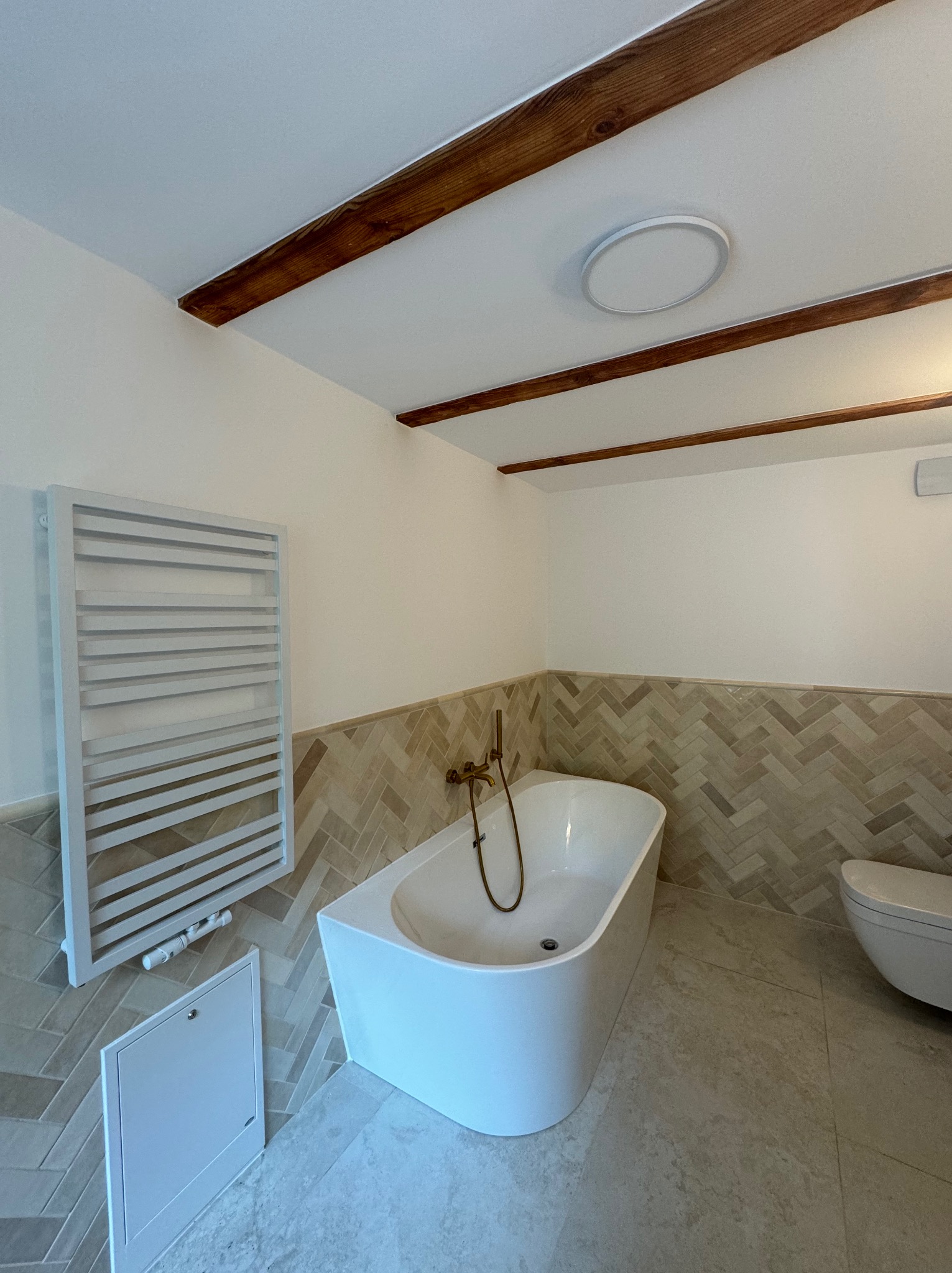 Biała, wolnostojąca wanna z mosiężną armaturą w nowoczesnej łazience z drewnianymi belkami na suficie i grzejnikiem drabinkowym. Ściany wyłożone płytkami w jodełkę.