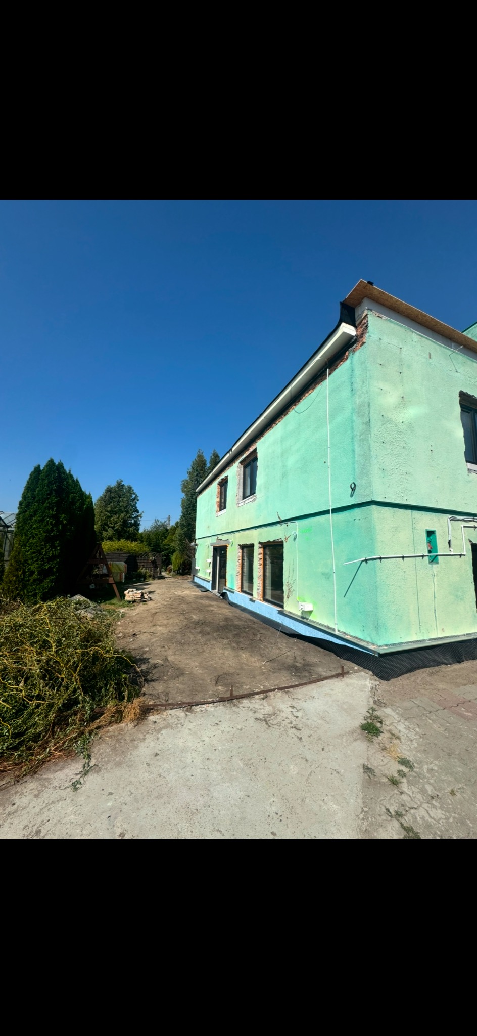 Dwupiętrowy budynek z zieloną elewacją w trakcie prac remontowych, widoczne niezamaskowane okna i fragmenty cegieł, zarośla i betonowy plac przed budynkiem, błękitne niebo w tle.