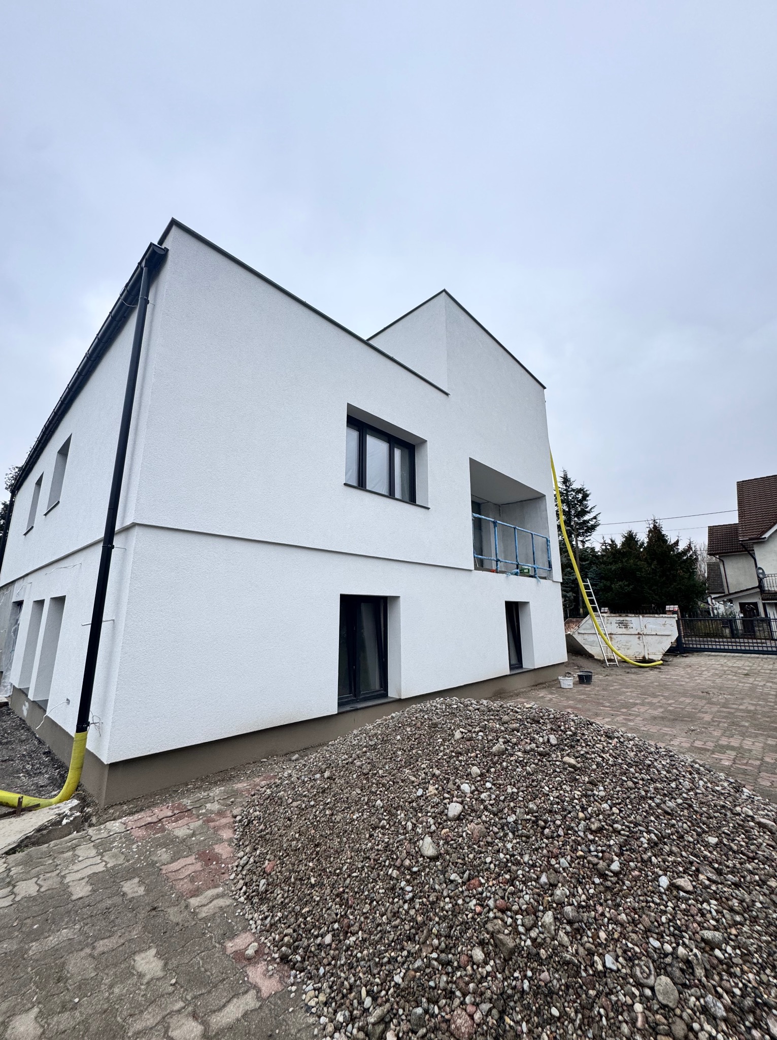 Nowoczesny, biały dom jednorodzinny z płaskim dachem w trakcie wykończenia elewacji, widoczna sterta kamieni na podjeździe i żółty wąż do podawania materiałów.
