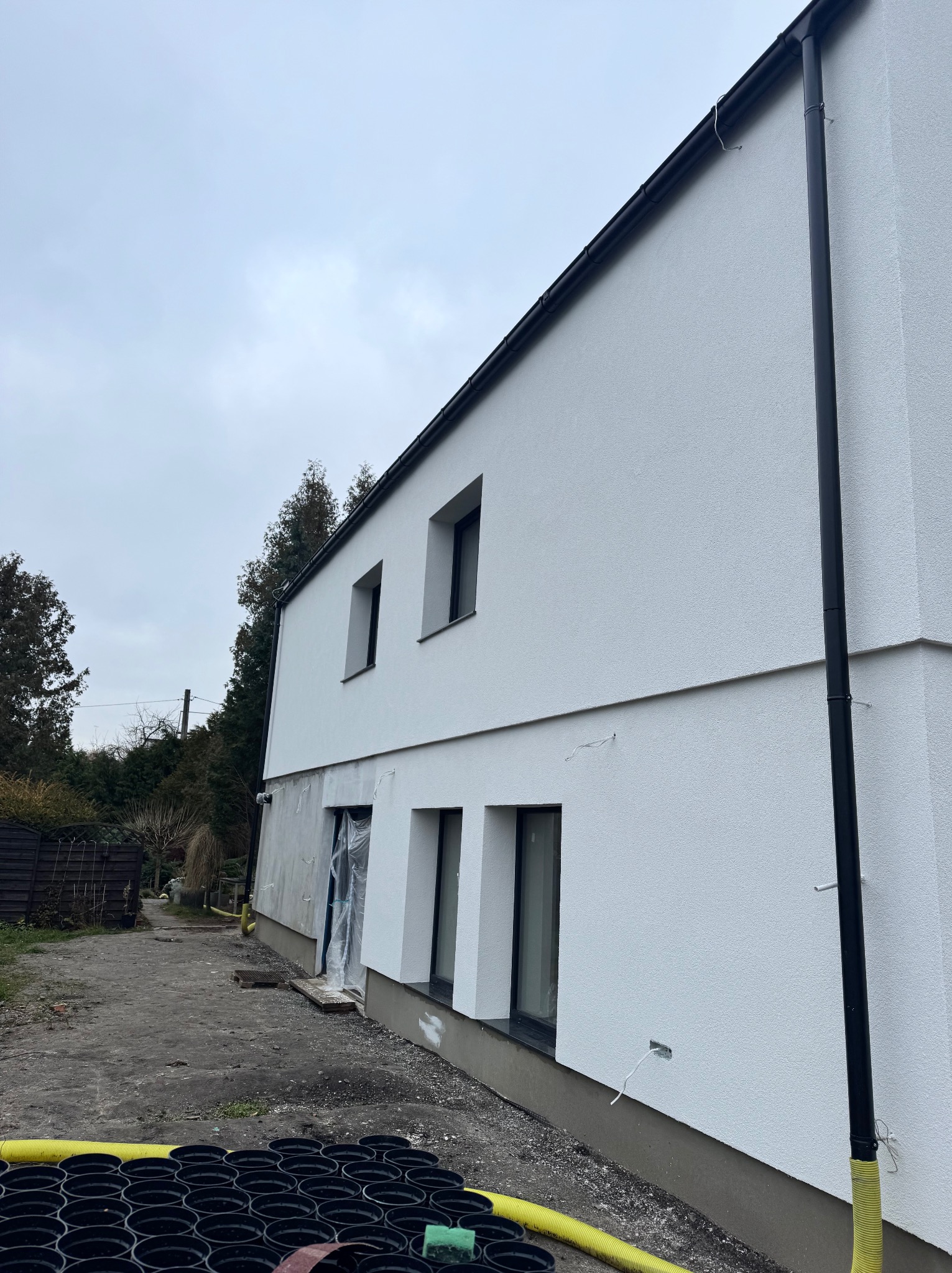 Elewacja budynku w trakcie prac wykończeniowych, widoczne okna, rynna, fragment ściany z niedokończonym tynkiem oraz czarne doniczki w tle.