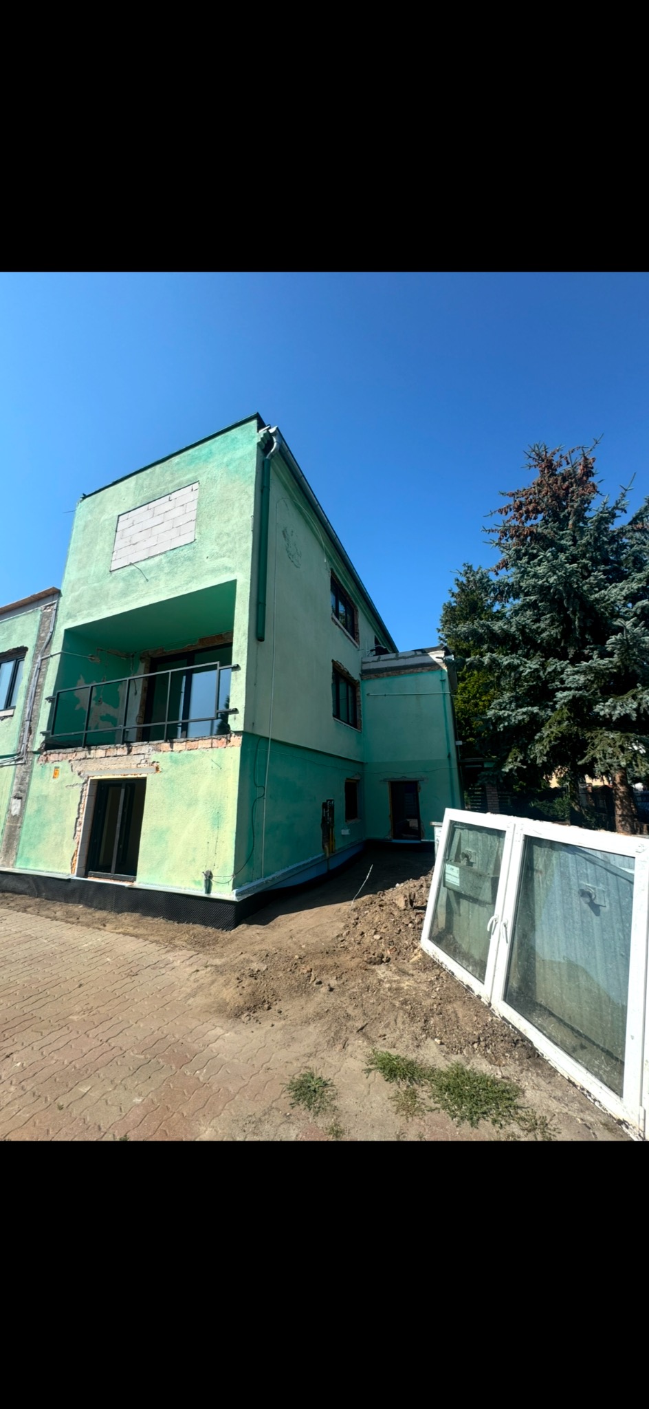 Budynek w trakcie prac remontowych z widocznymi brakami w elewacji, nowymi oknami na balkonie, zabezpieczeniem fundamentów i oknami czekającymi na montaż.