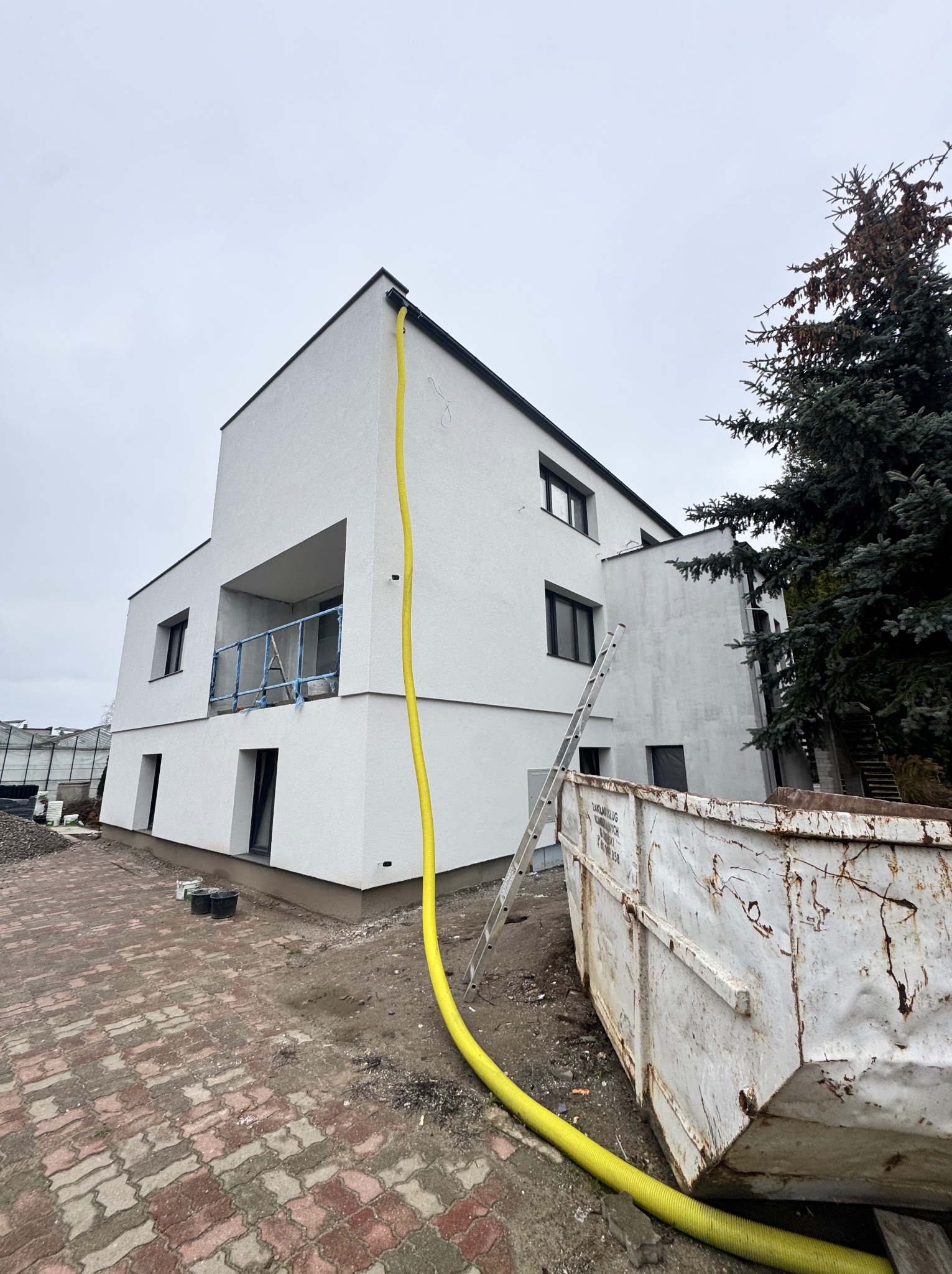 Biały, dwupiętrowy budynek w trakcie wykończenia, z żółtym wężem spustowym przymocowanym do dachu, stalową drabiną opartą o ścianę i kontenerem na gruz obok, na tle pochmurnego nieba.
