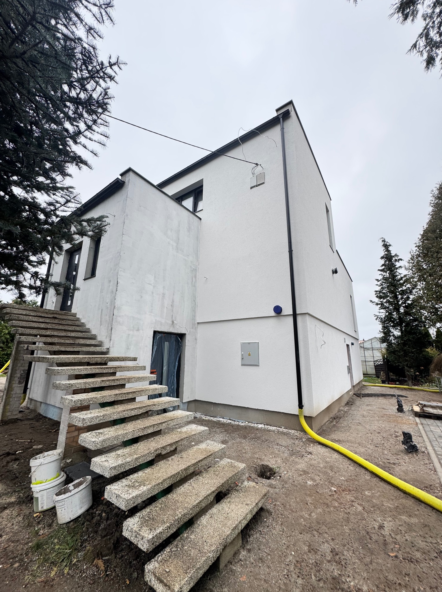 Nowoczesny dom z białym tynkiem zewnętrznym w trakcie wykończenia, widoczne betonowe schody wejściowe, przygotowany teren wokół budynku i żółty wąż ogrodowy.