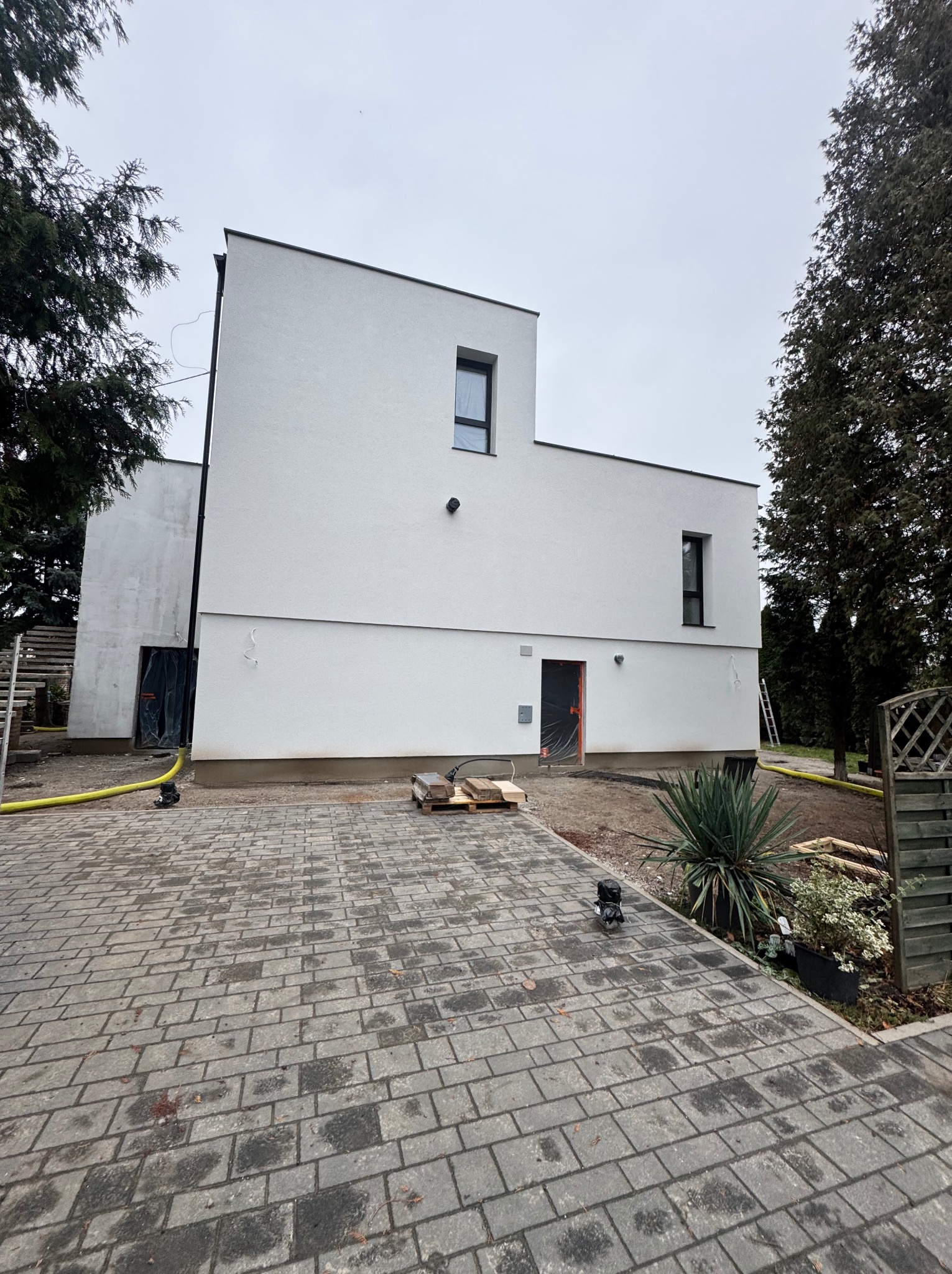 Dwukondygnacyjny, biały budynek z płaskim dachem w stylu modernistycznym, z widocznymi śladami prac wykończeniowych na elewacji i otoczeniu, w tym żółty wąż i palety z materiałami budowlanymi.