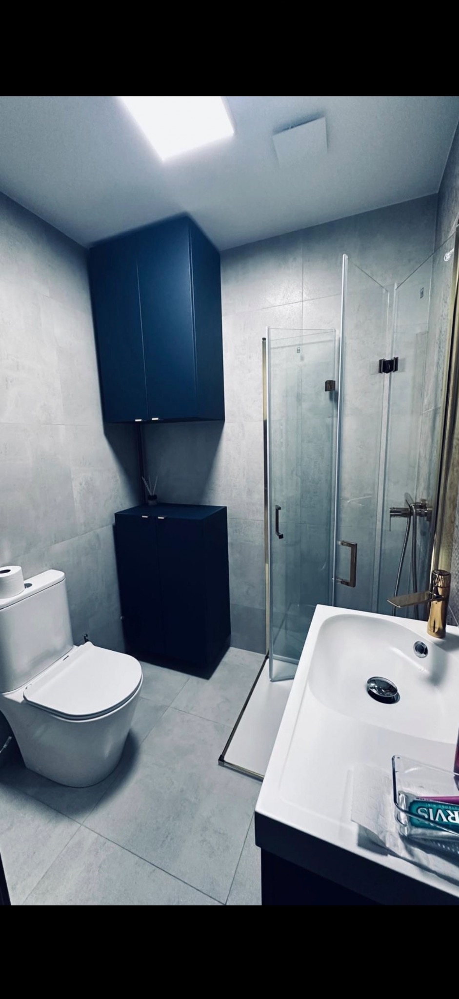 Nowoczesna łazienka z szarymi płytkami, kabiną prysznicową ze złotymi akcentami i ciemnoniebieskimi szafkami. Umywalka z blatem i toaleta w minimalistycznym stylu.