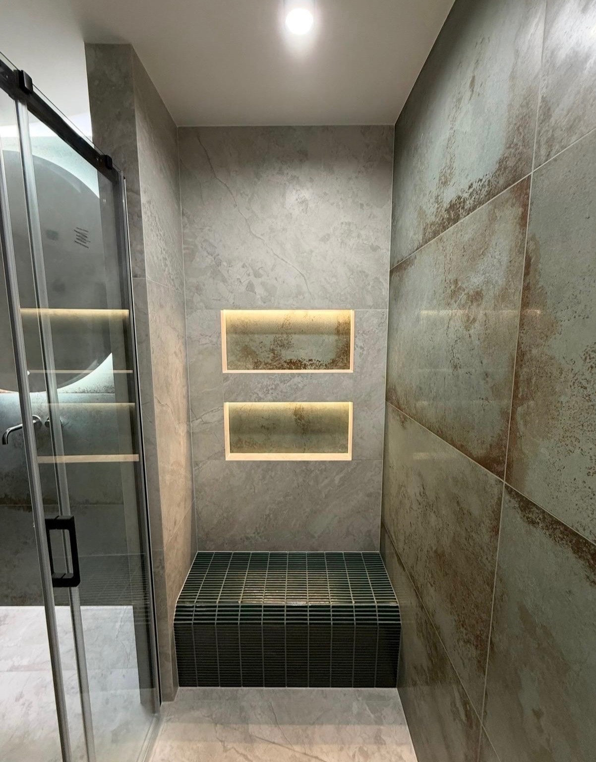 Nowoczesna łazienka z kabiną prysznicową, siedziskiem z mozaiki i podświetlanymi wnękami w szarej, marmurkowej okładzinie ściennej. Minimalistyczny design.