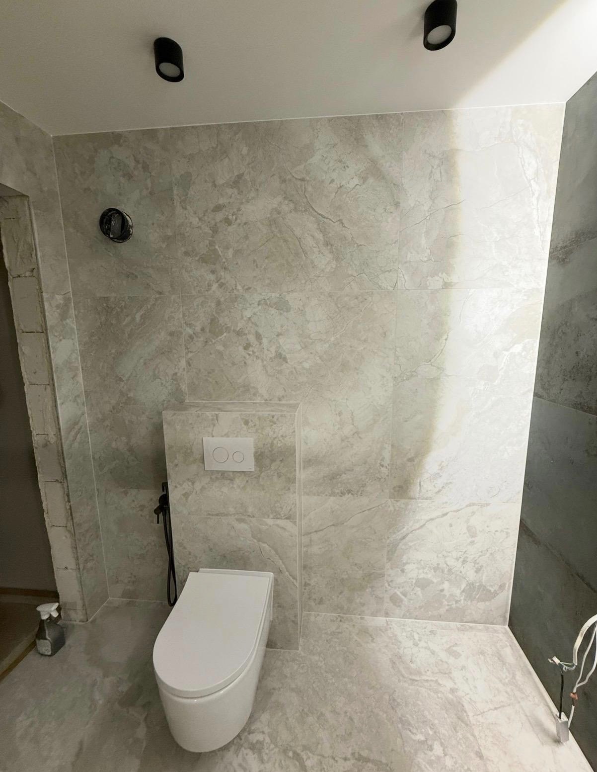 Wykończona łazienka z beżowymi płytkami imitującymi marmur, wiszącą toaletą i czarnymi punktowymi lampami sufitowymi. Widoczne przyłącza i fragmenty niezakończonych prac.