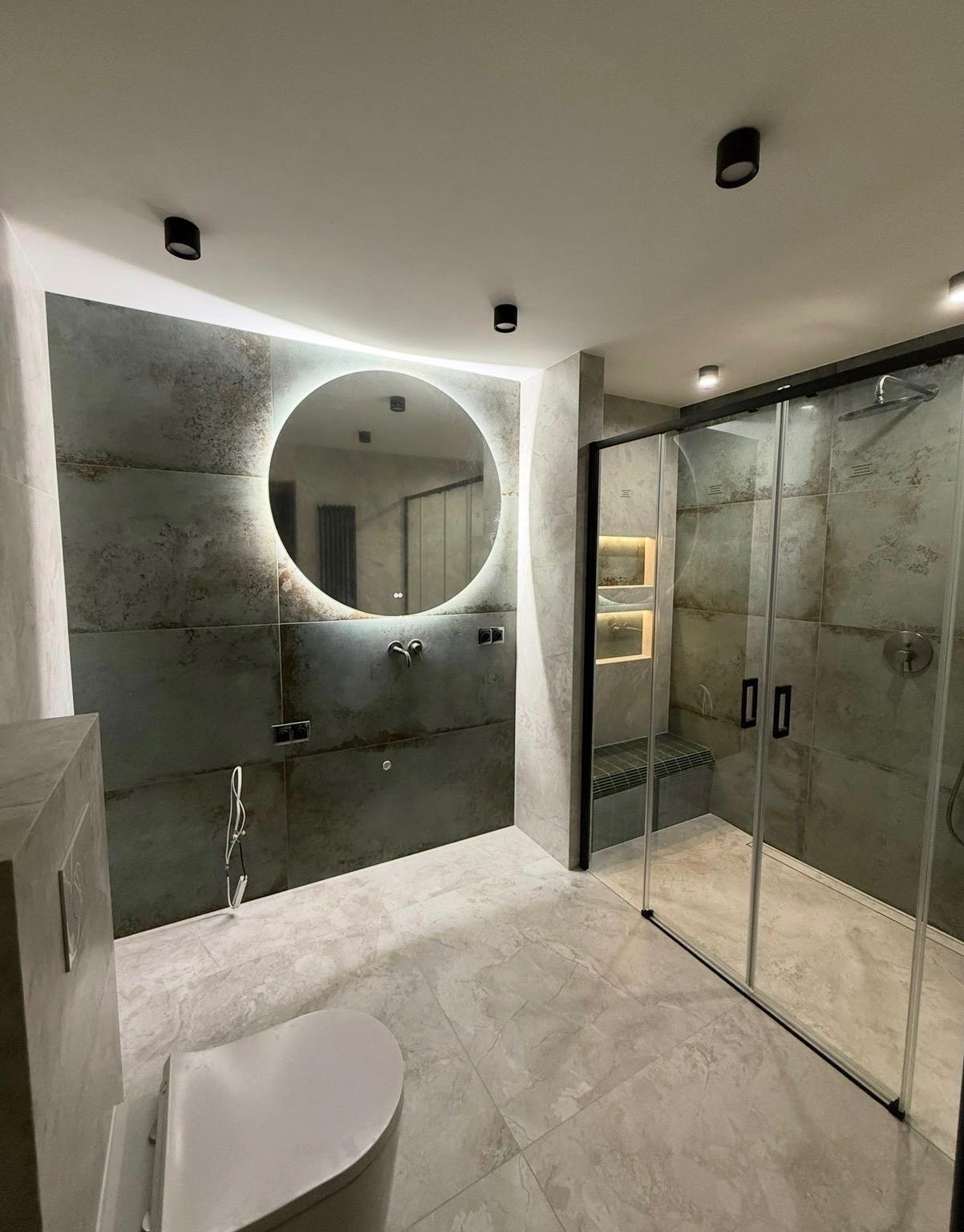 Nowoczesna łazienka z kabiną prysznicową typu walk-in, okrągłym lustrem z podświetleniem LED i płytkami imitującymi beton. Minimalistyczny design z czarnymi akcentami.