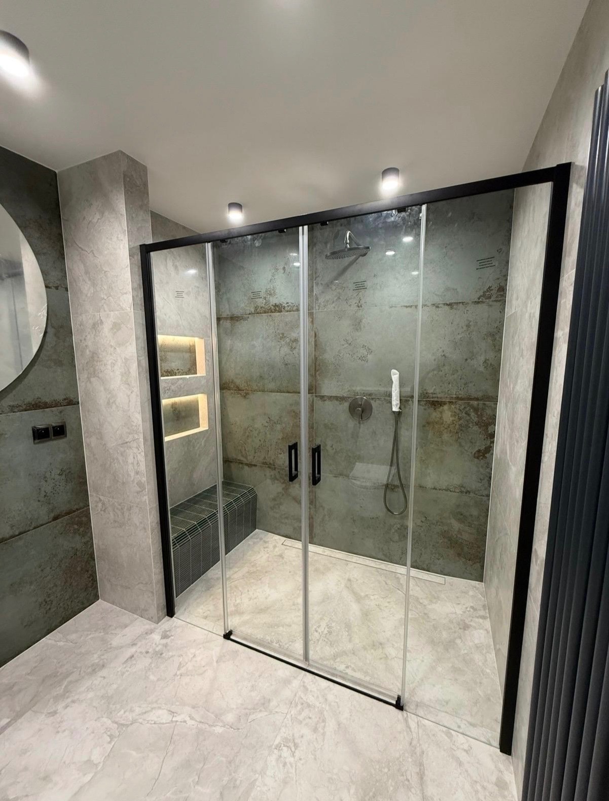 Nowoczesna kabina prysznicowa z czarnymi ramami i szklanymi drzwiami przesuwnymi, w łazience wykończonej płytkami imitującymi beton i marmurową podłogą.