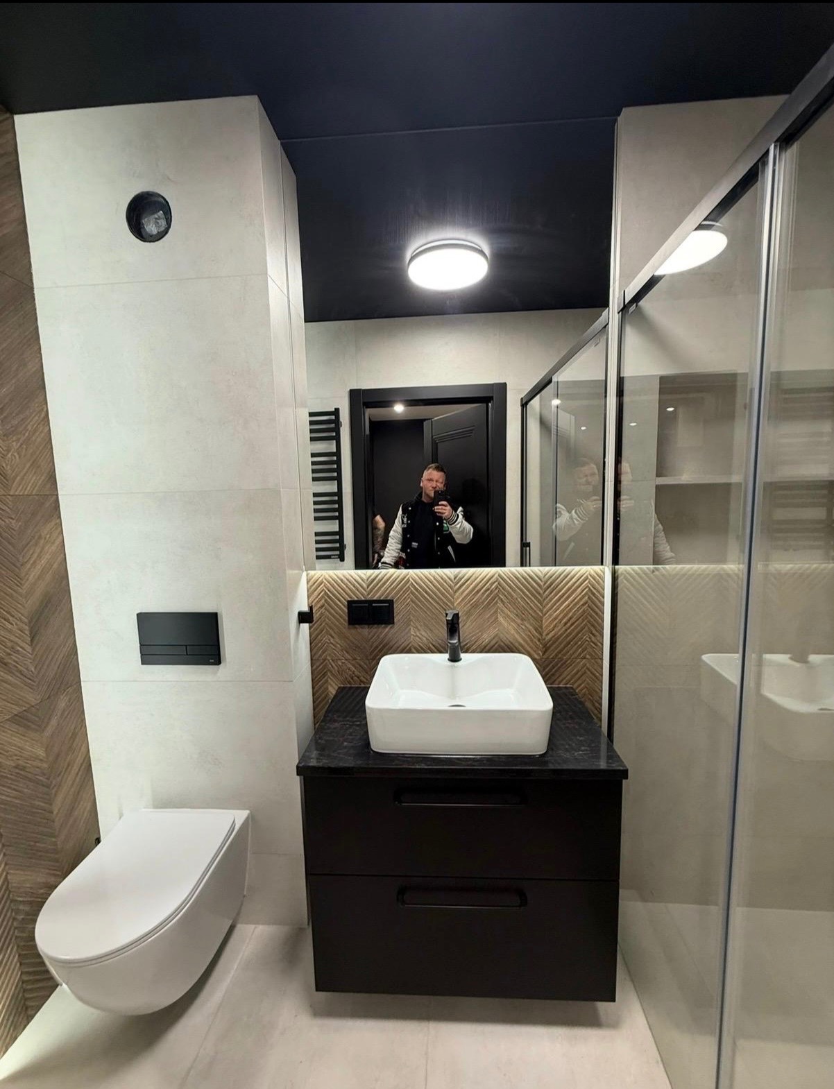 Nowoczesna łazienka z białą ceramiką, czarnymi akcentami i drewnianą mozaiką na ścianie, odbicie osoby robiącej zdjęcie w lustrze.