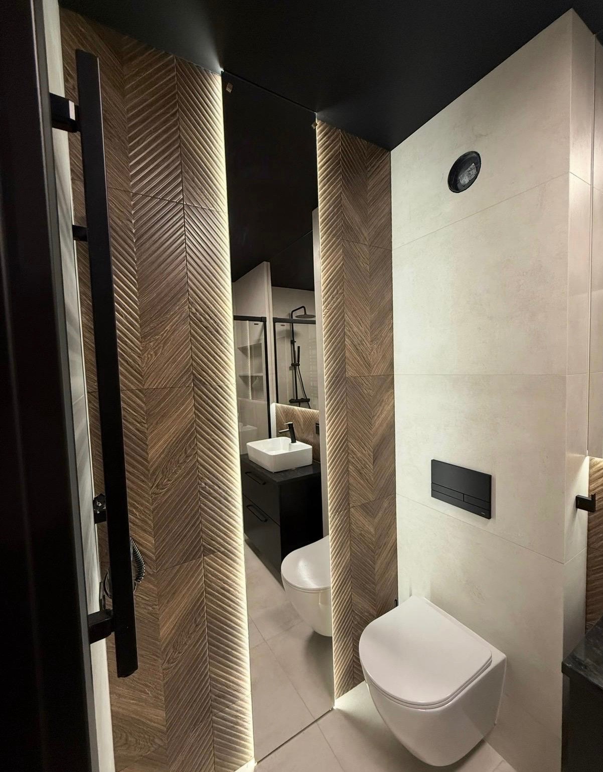 Nowoczesna łazienka z drewnopodobnymi płytkami ułożonymi w jodełkę, podświetlanym lustrem i minimalistyczną armaturą. Widok na kabinę prysznicową w odbiciu.