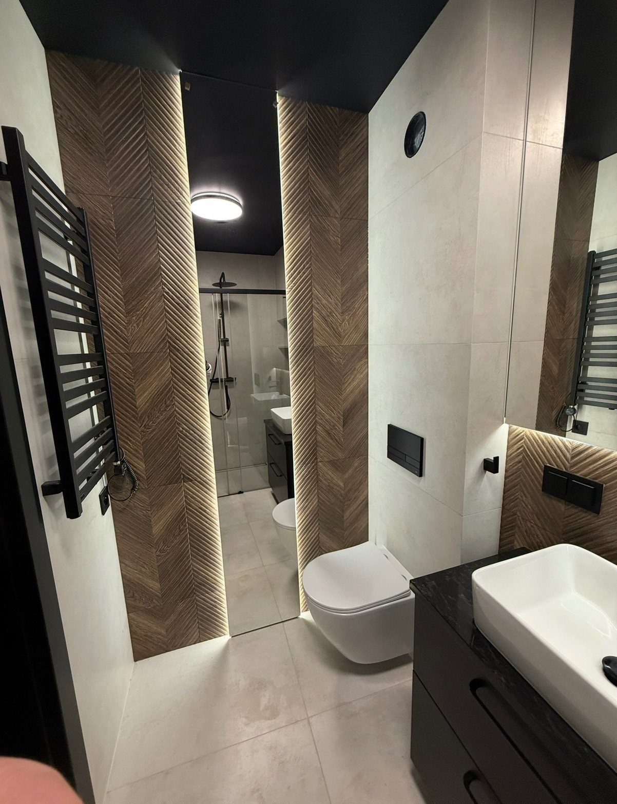 Nowoczesna łazienka z drewnopodobnymi płytkami ułożonymi w jodełkę, podświetlanymi lustrami i czarnymi akcentami, w tym grzejnikiem i armaturą.