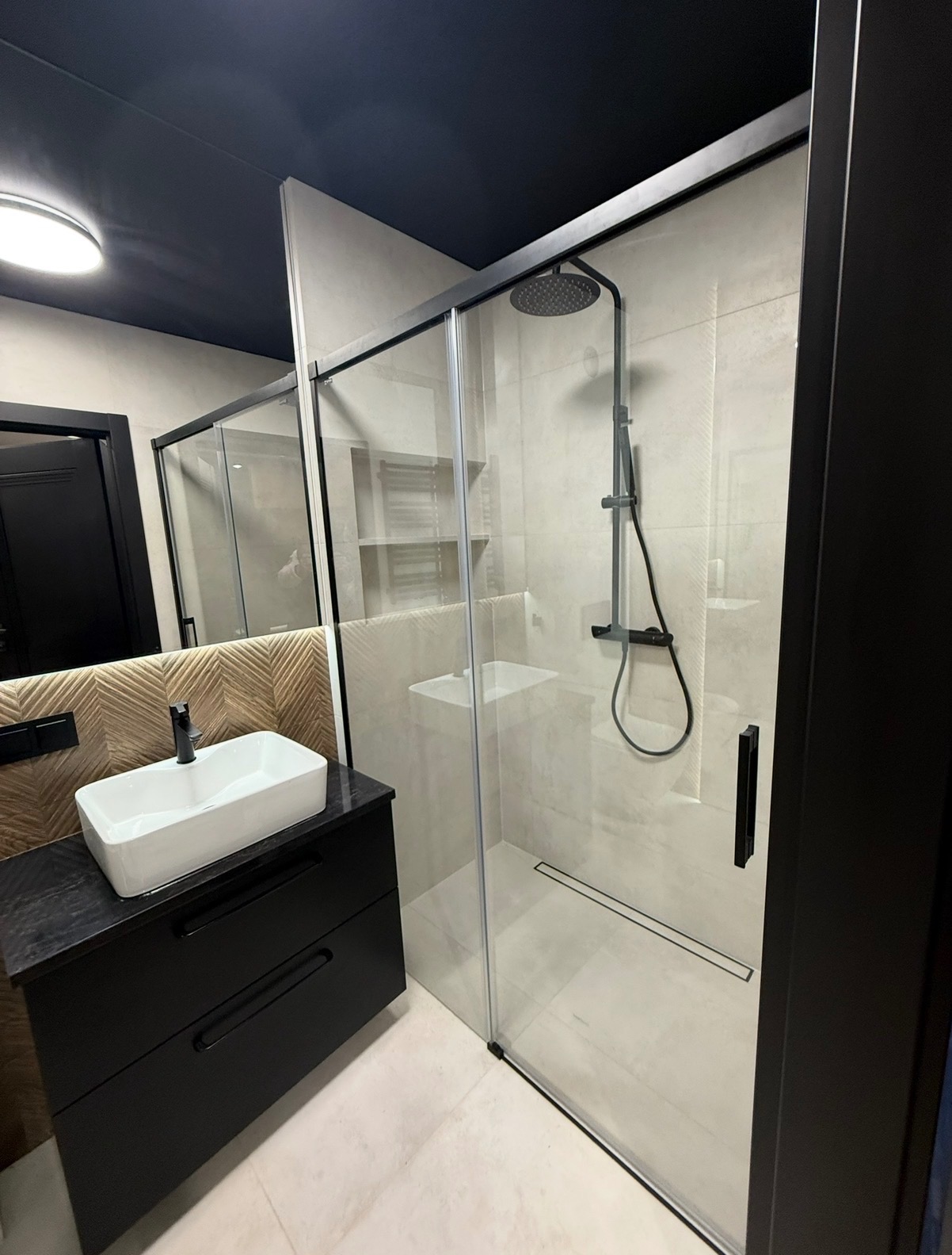 Nowoczesna łazienka z kabiną prysznicową typu walk-in, czarną armaturą i szafką pod umywalkę z blatem z ciemnego kamienia. Uwagę zwraca sufit w kolorze antracytu.