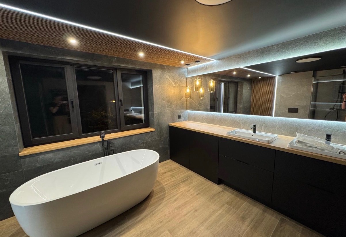 Nowoczesna łazienka z wolnostojącą wanną, czarnymi szafkami pod umywalką z drewnianym blatem i dużym lustrem z podświetleniem LED. Szare płytki na ścianach i drewnopodobna podłoga.