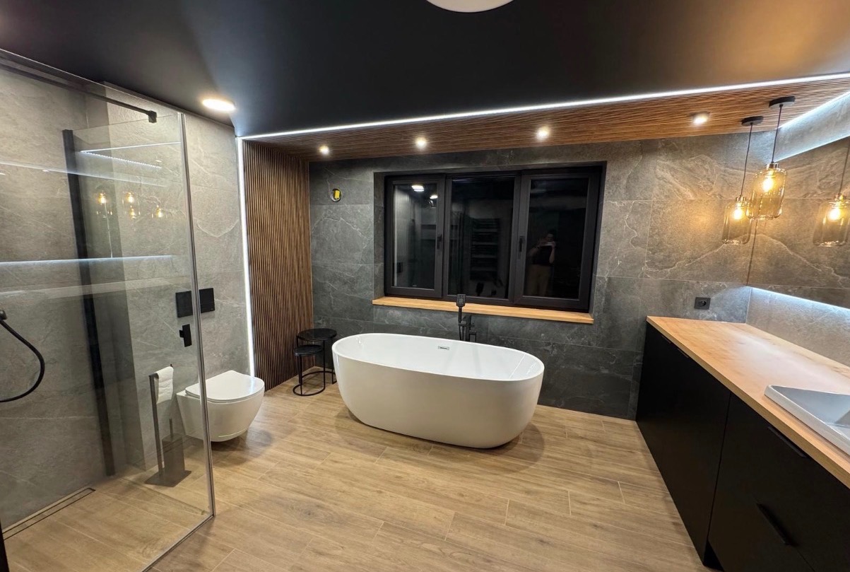 Nowoczesna łazienka z wolnostojącą wanną, kabiną prysznicową ze szklaną ścianą i drewnianymi akcentami na ścianie i pod sufitem. Uwagę przykuwają lampy wiszące.