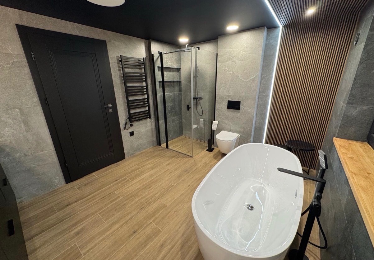 Nowoczesna łazienka z wolnostojącą wanną, kabiną prysznicową typu walk-in i drewnianymi panelami ściennymi. Uwagę zwraca czarna armatura i oświetlenie LED.