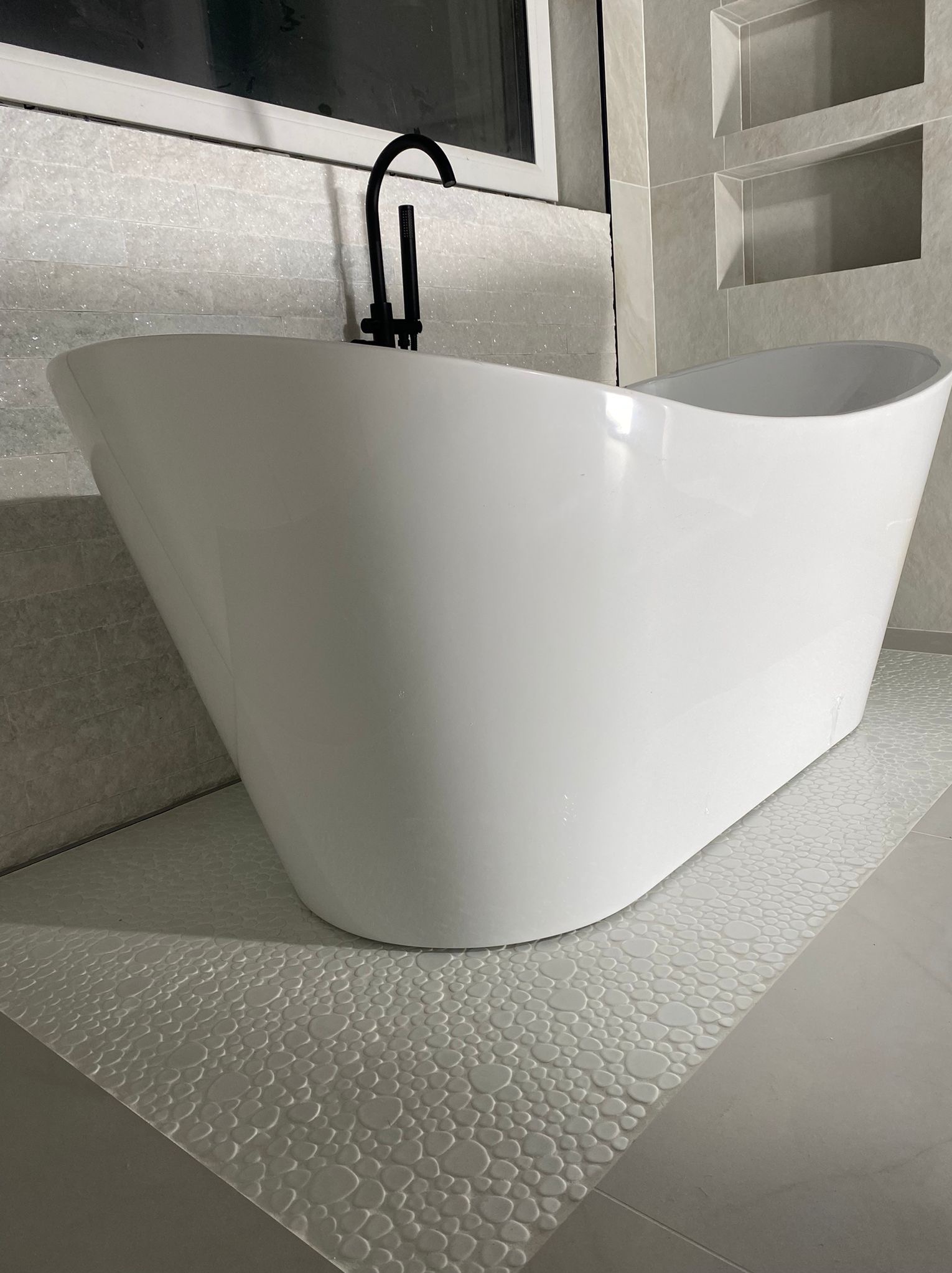 Nowoczesna, wolnostojąca wanna w łazience z szarymi płytkami i czarną armaturą. Dekoracyjne płytki pod wanną imitują kamienny dywan.