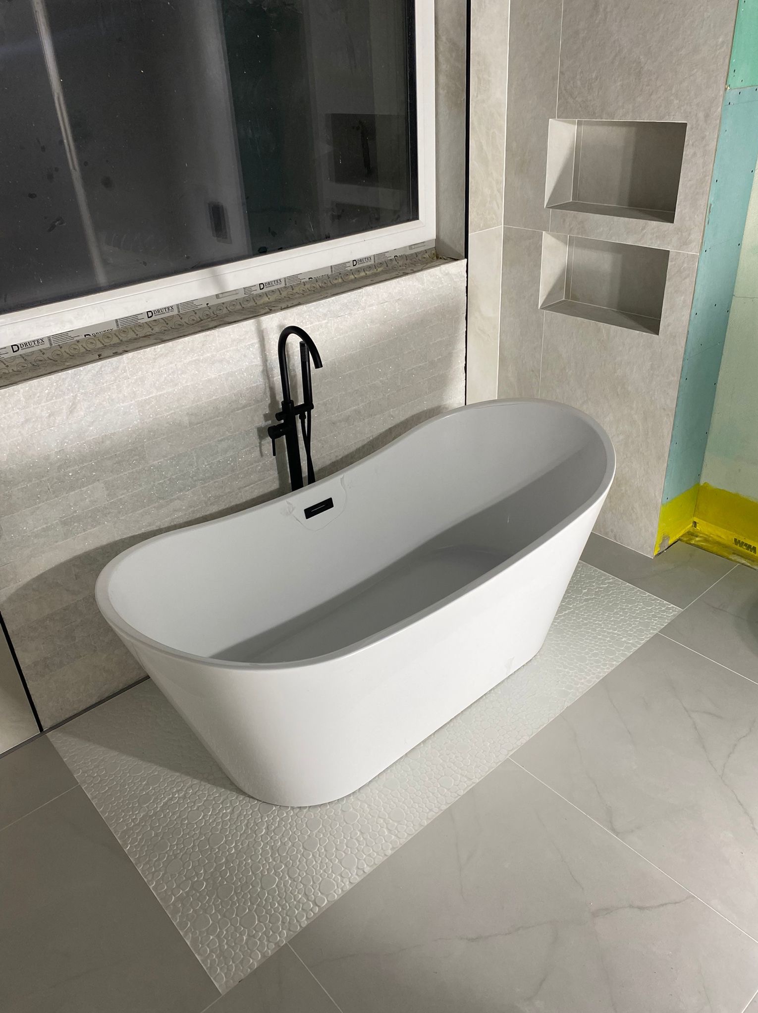 Nowoczesna, biała wanna wolnostojąca z czarną armaturą na tle szarych płytek i wnęk w ścianie. Widoczne okno i fragment ściany z płyt karton-gips.