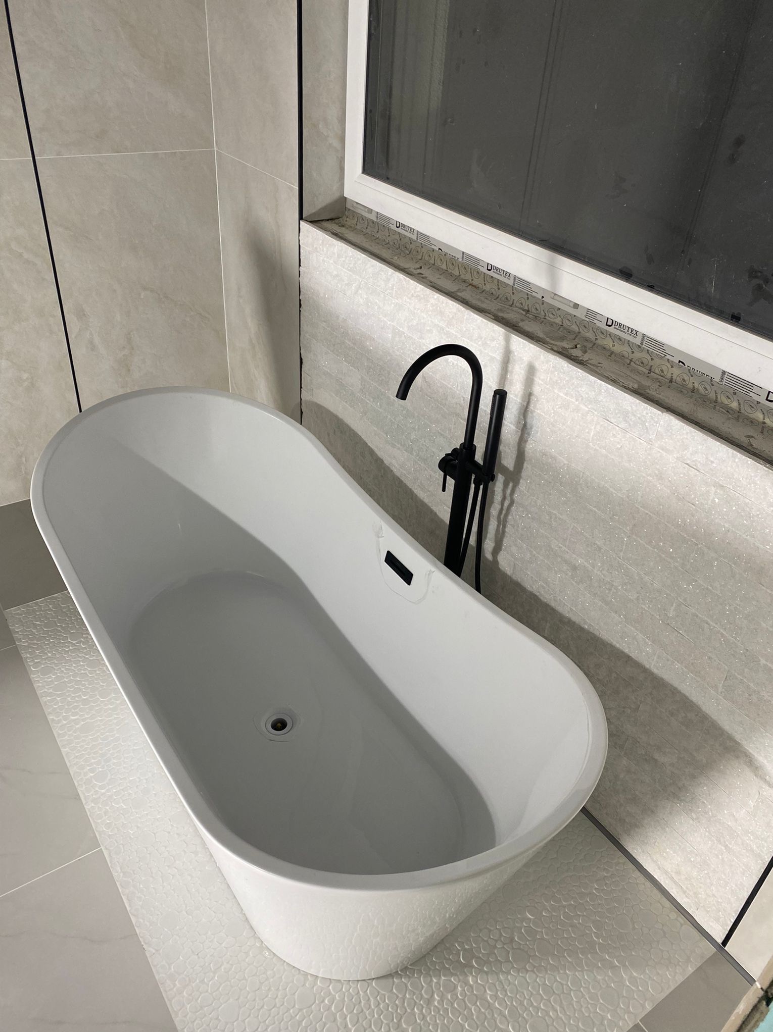 Biała wanna wolnostojąca z czarną armaturą na tle jasnych płytek w łazience z oknem. Minimalistyczny design, nowoczesne wykończenie.