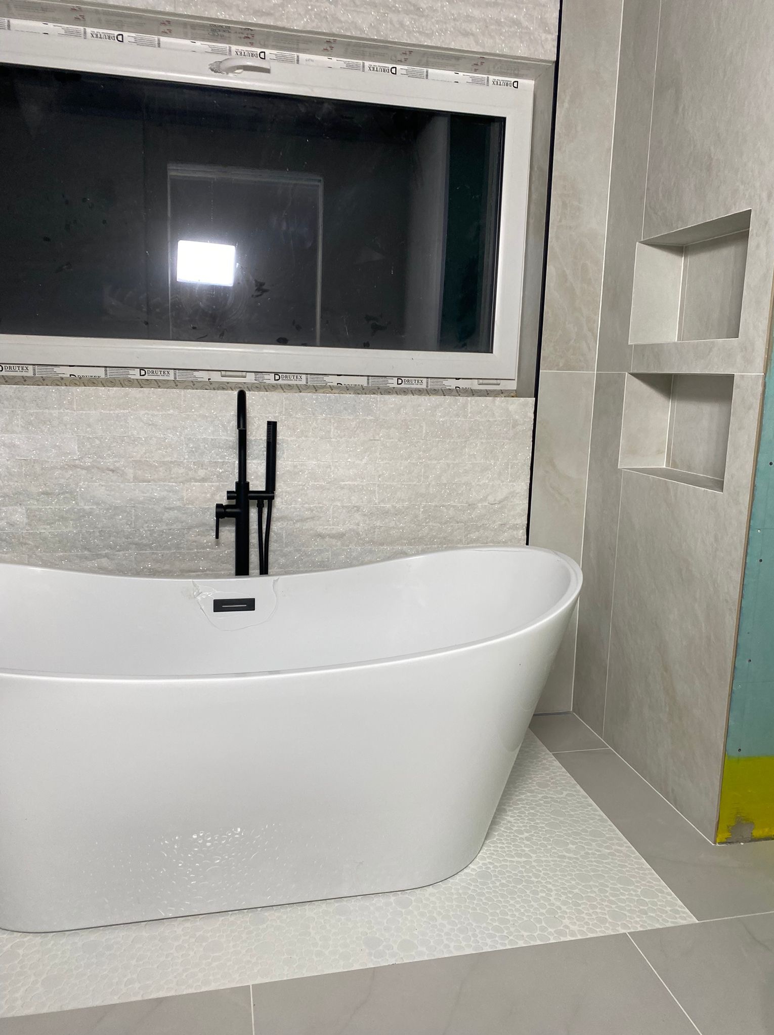 Nowoczesna łazienka z wolnostojącą białą wanną, czarną armaturą i jasnymi płytkami imitującymi kamień. Widoczne wnęki w ścianie i okno.