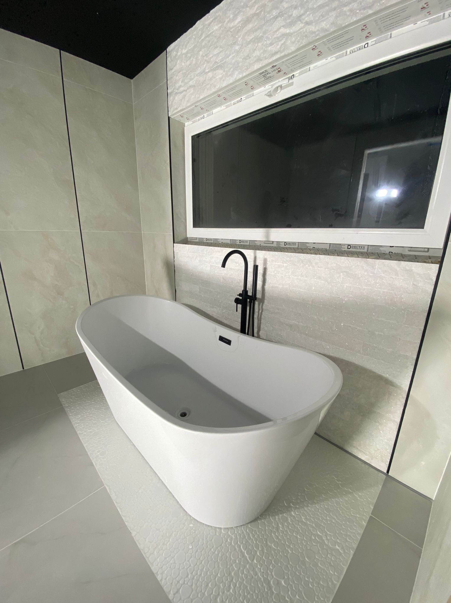 Nowoczesna, wolnostojąca wanna w jasnej łazience z oknem. Uwagę przykuwa czarna bateria i dekoracyjne płytki imitujące kamień na ścianie pod oknem.