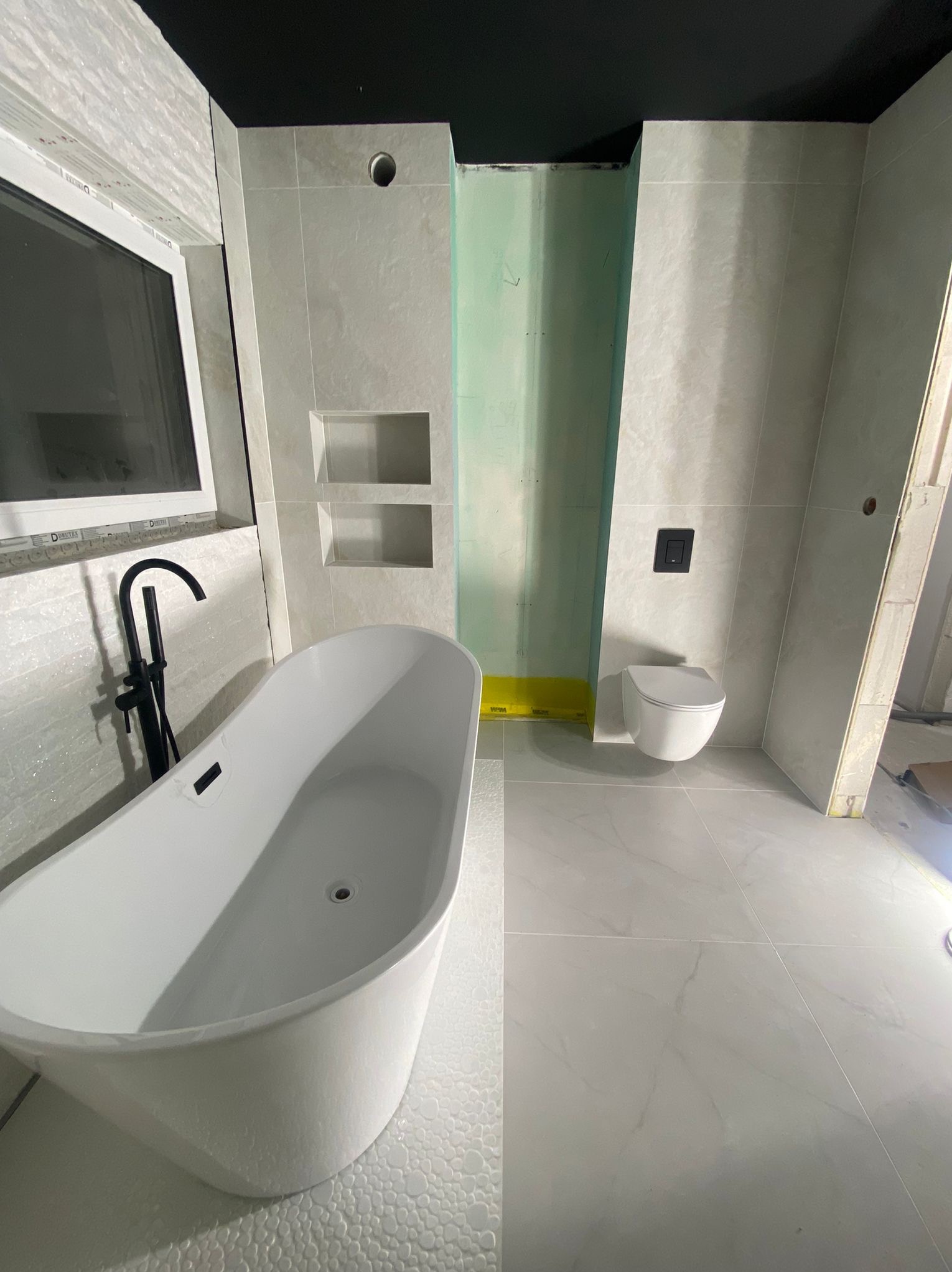 Nowoczesna łazienka w trakcie wykańczania: biała wanna wolnostojąca, podwieszana toaleta, jasne płytki imitujące marmur, czarna armatura i sufit.