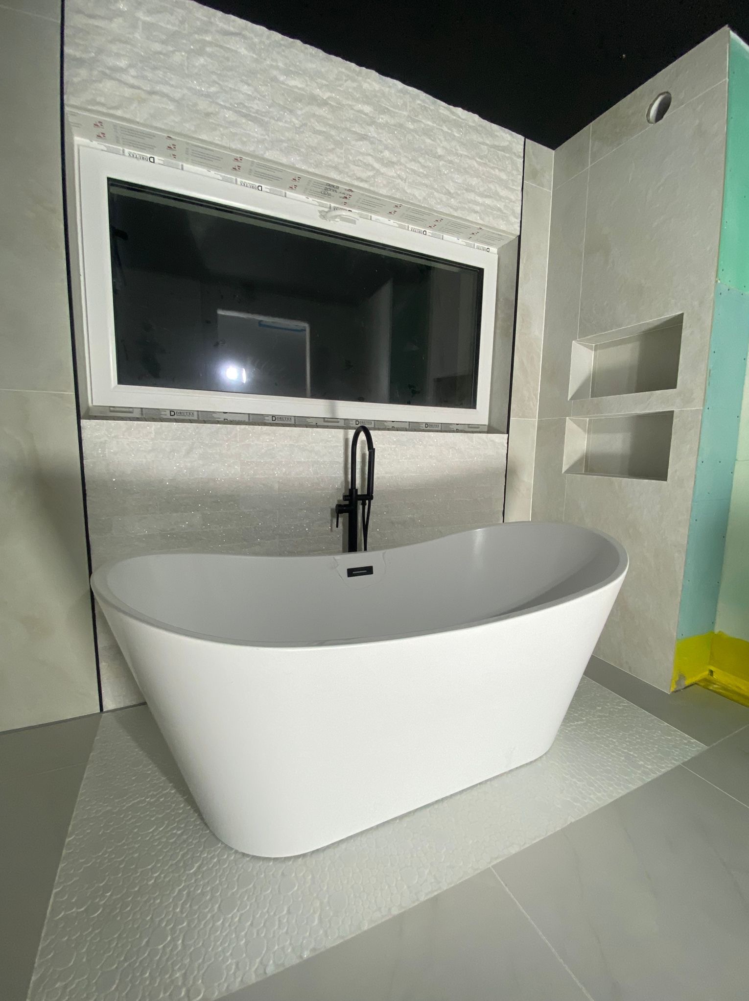 Nowoczesna, wolnostojąca wanna z czarną armaturą na tle wykończonej płytkami łazienki z oknem i wnękami. Widoczna płyta karton-gips na ścianie.