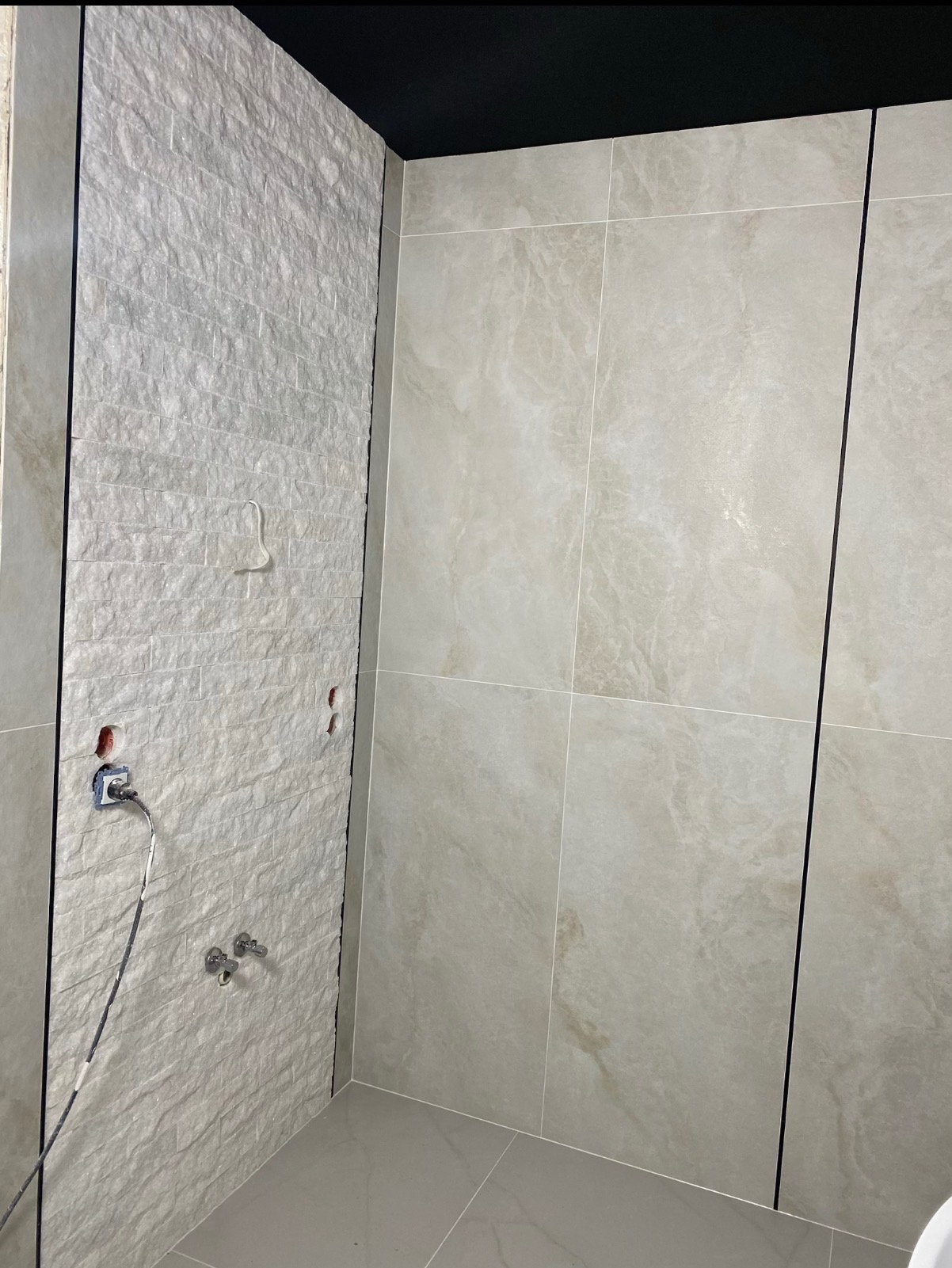 Wykończona łazienka z beżowymi płytkami imitującymi marmur i szarą ścianą z fakturą kamienia. Widoczne przyłącza wodne i gniazdko elektryczne. Czarny sufit.