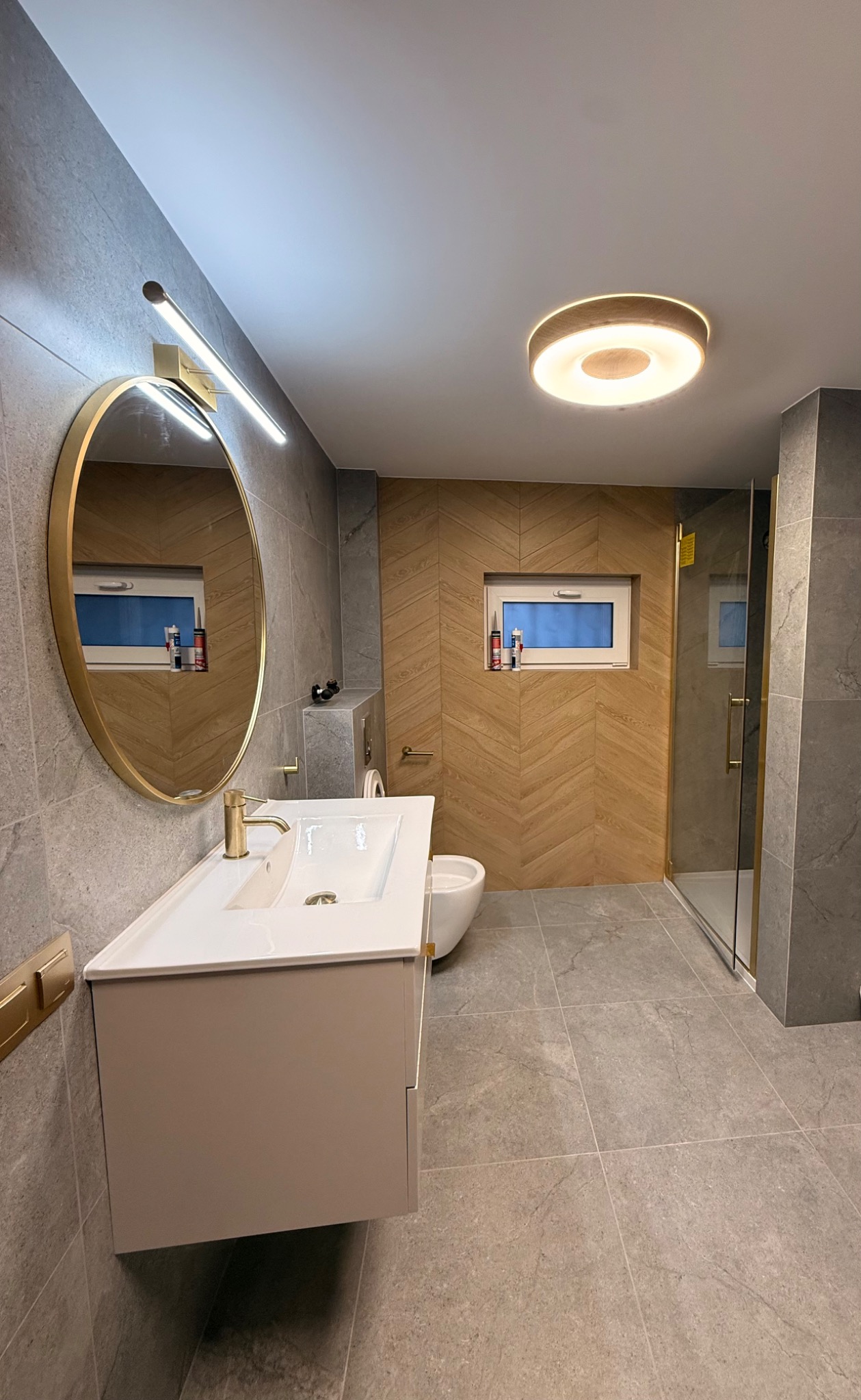 Nowoczesna łazienka w Gorzowie Wielkopolskim z szarymi płytkami, drewnianą ścianą w jodełkę i okrągłym lustrem. Umywalka z szafką i kabina prysznicowa.