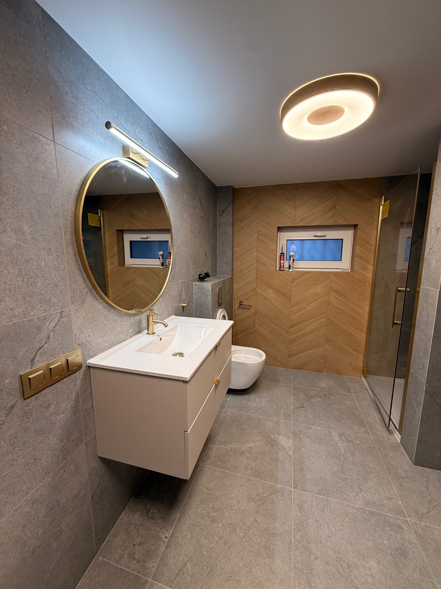 Nowoczesna łazienka z szarymi płytkami, okrągłym lustrem w złotej ramie, drewnianą ścianą w jodełkę i kabiną prysznicową ze złotymi detalami.