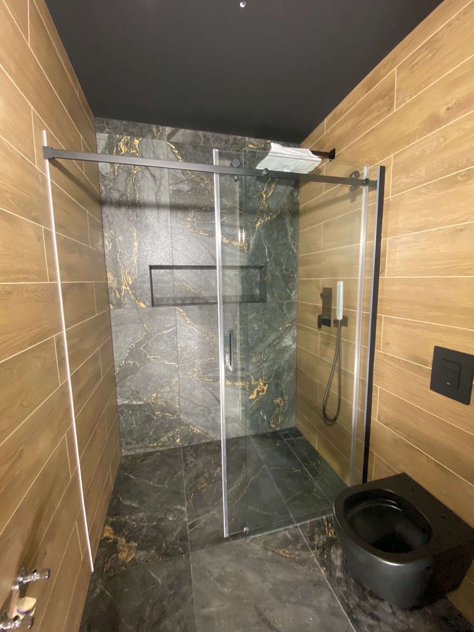 Nowoczesna łazienka z kabiną prysznicową typu walk-in, wykończona płytkami imitującymi drewno i marmur z złotymi żyłami, czarna armatura i podwieszana toaleta.