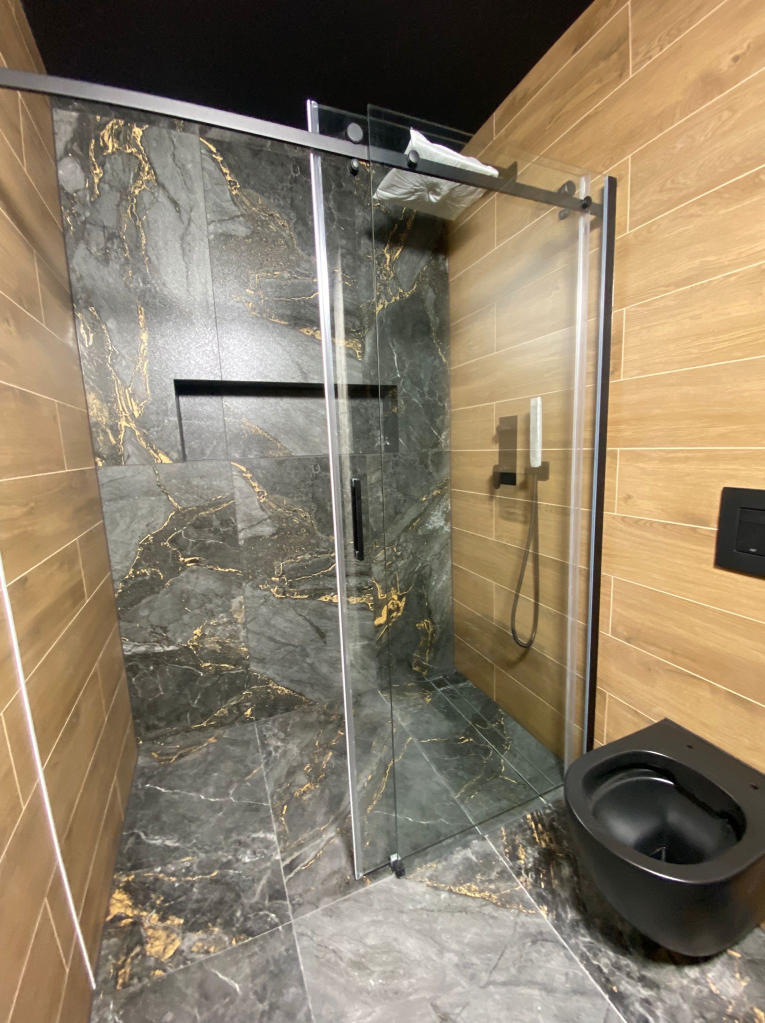 Nowoczesna łazienka po remoncie: kabina prysznicowa ze złotymi żyłami na szarych płytkach, drewnopodobne płytki na ścianach, czarna wisząca toaleta.