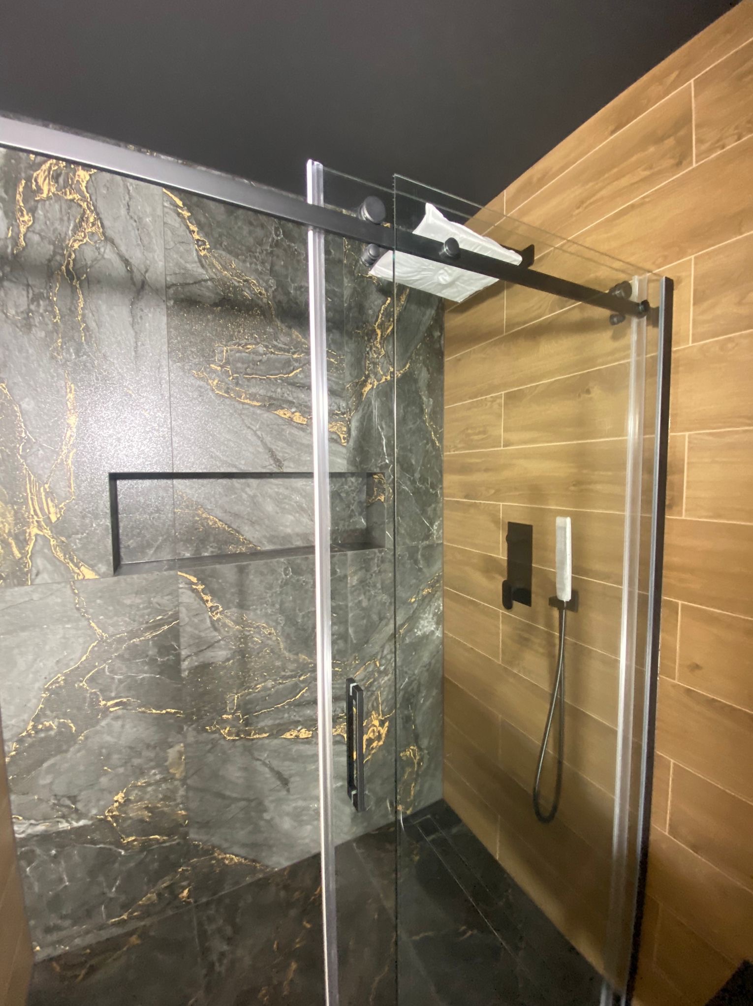 Nowoczesna kabina prysznicowa z przesuwnymi drzwiami, wykończona płytkami imitującymi marmur z złotymi żyłami i drewnopodobnymi, z czarną armaturą i sufitem.