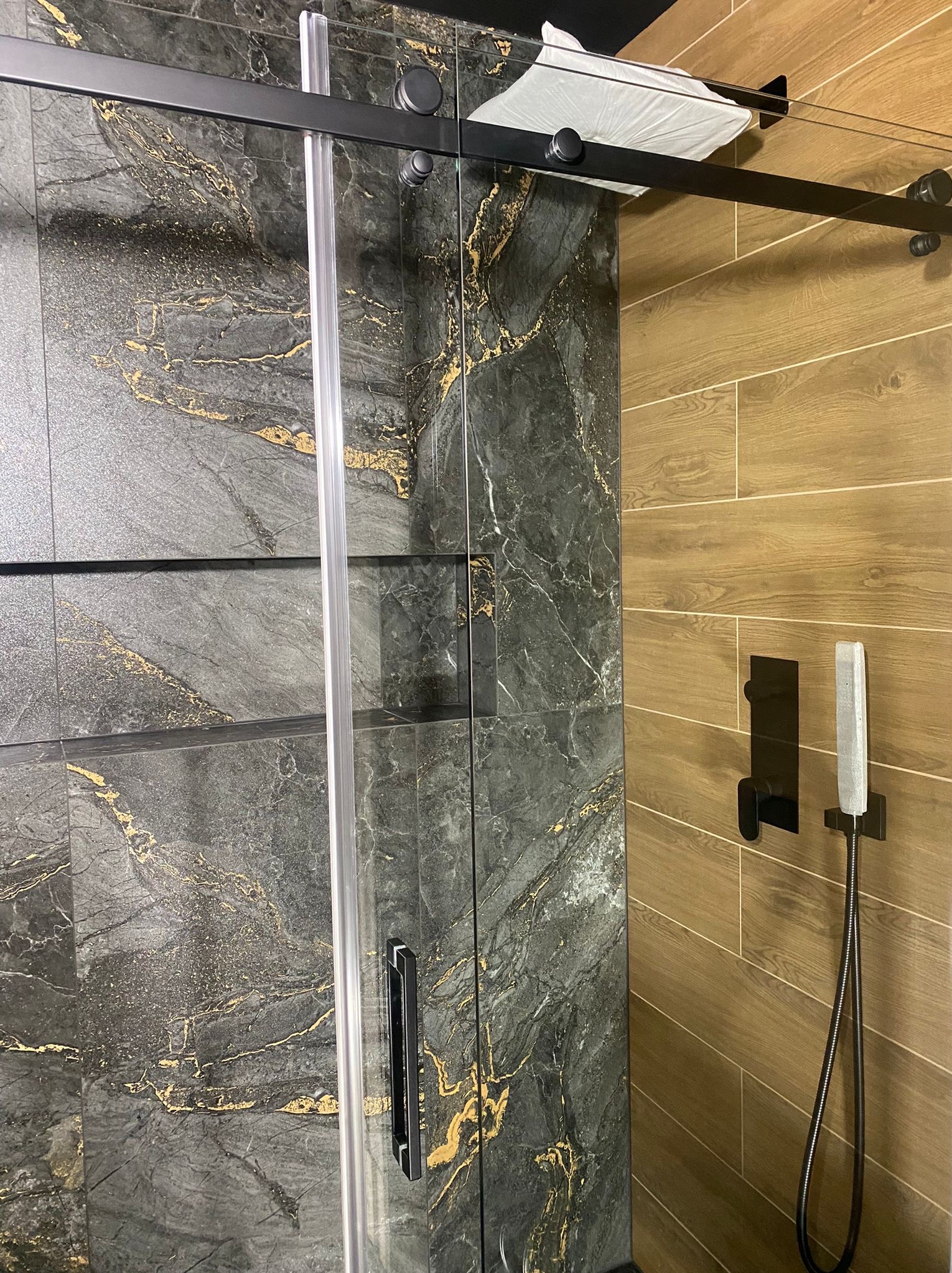 Kabina prysznicowa z przesuwnymi drzwiami, wykończona płytkami imitującymi marmur z złotymi żyłami i drewnopodobnymi panelami. Nowoczesny design, czarne akcenty.