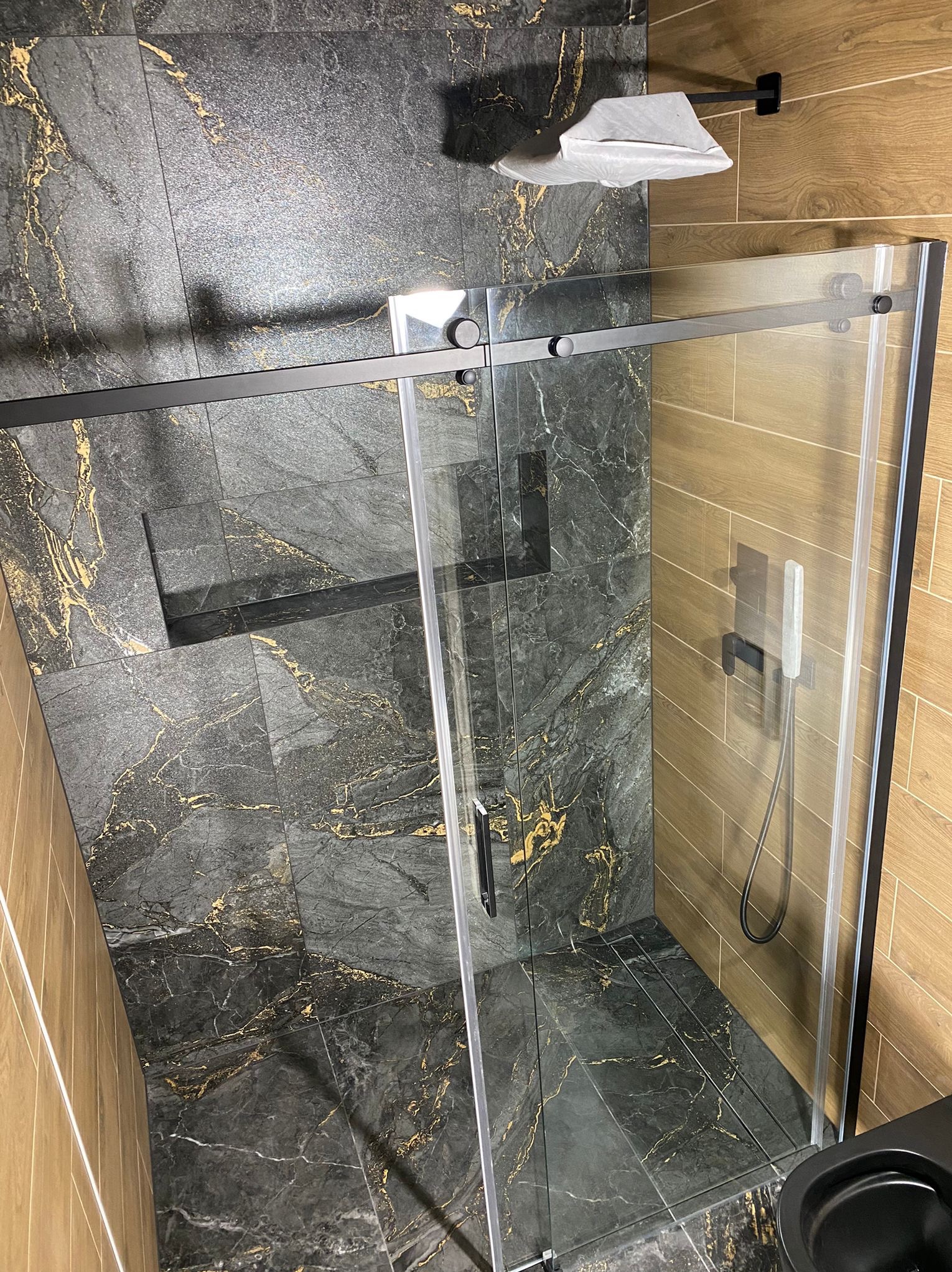 Nowoczesna kabina prysznicowa z czarnymi profilami i marmurowymi płytkami w odcieniach szarości i złota. Widok z góry, akcent na detale wykończenia.