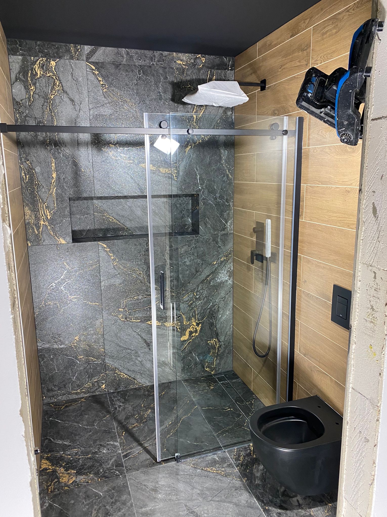 Nowoczesna łazienka po remoncie: kabina prysznicowa ze szklanymi drzwiami przesuwnymi, czarna armatura i płytki imitujące marmur z złotymi żyłami.