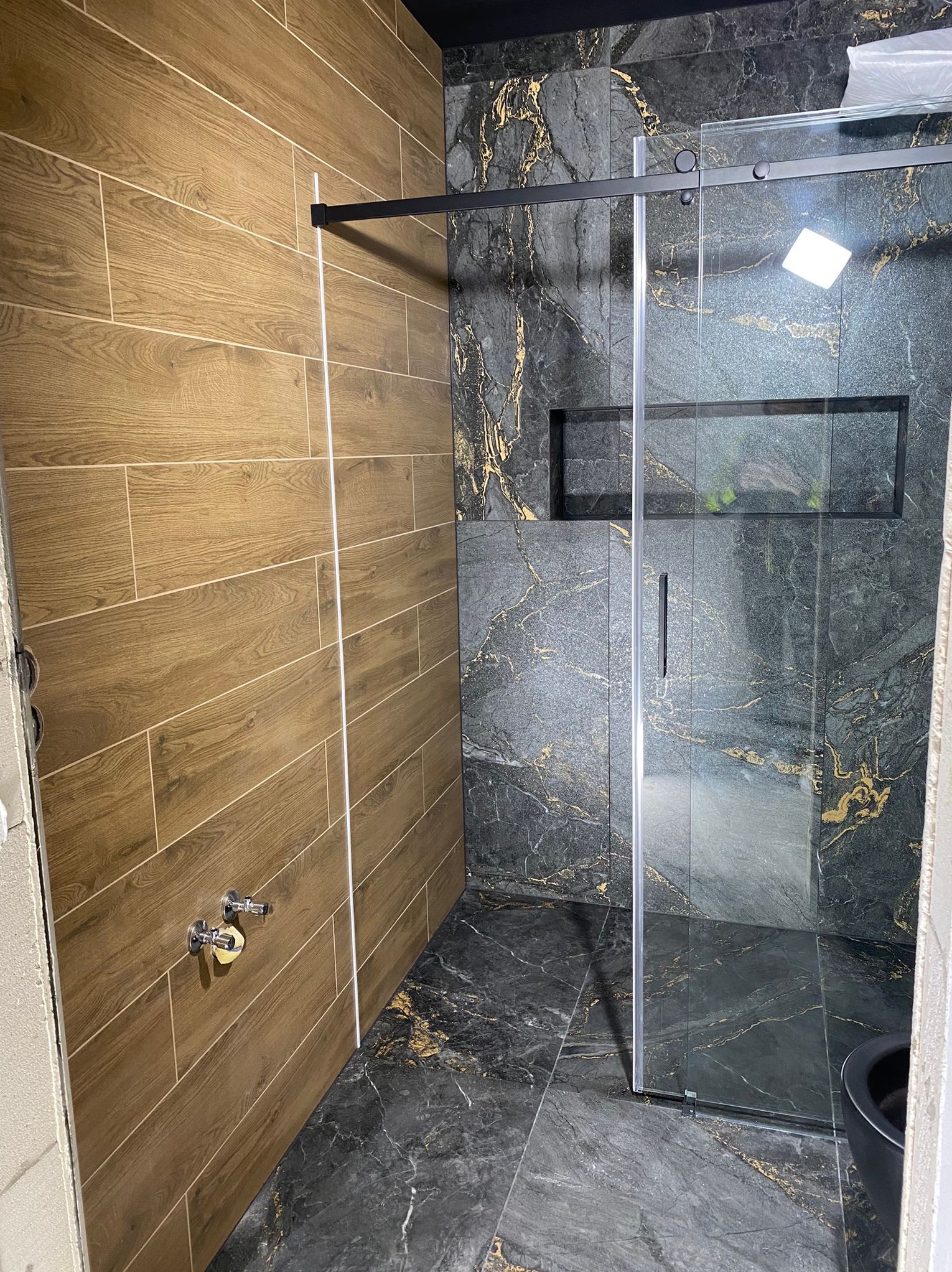 Nowoczesna łazienka po remoncie: drewnopodobne płytki na ścianie, kabina prysznicowa ze szkła i czarnych profili, marmurowe płytki z złotymi żyłami.