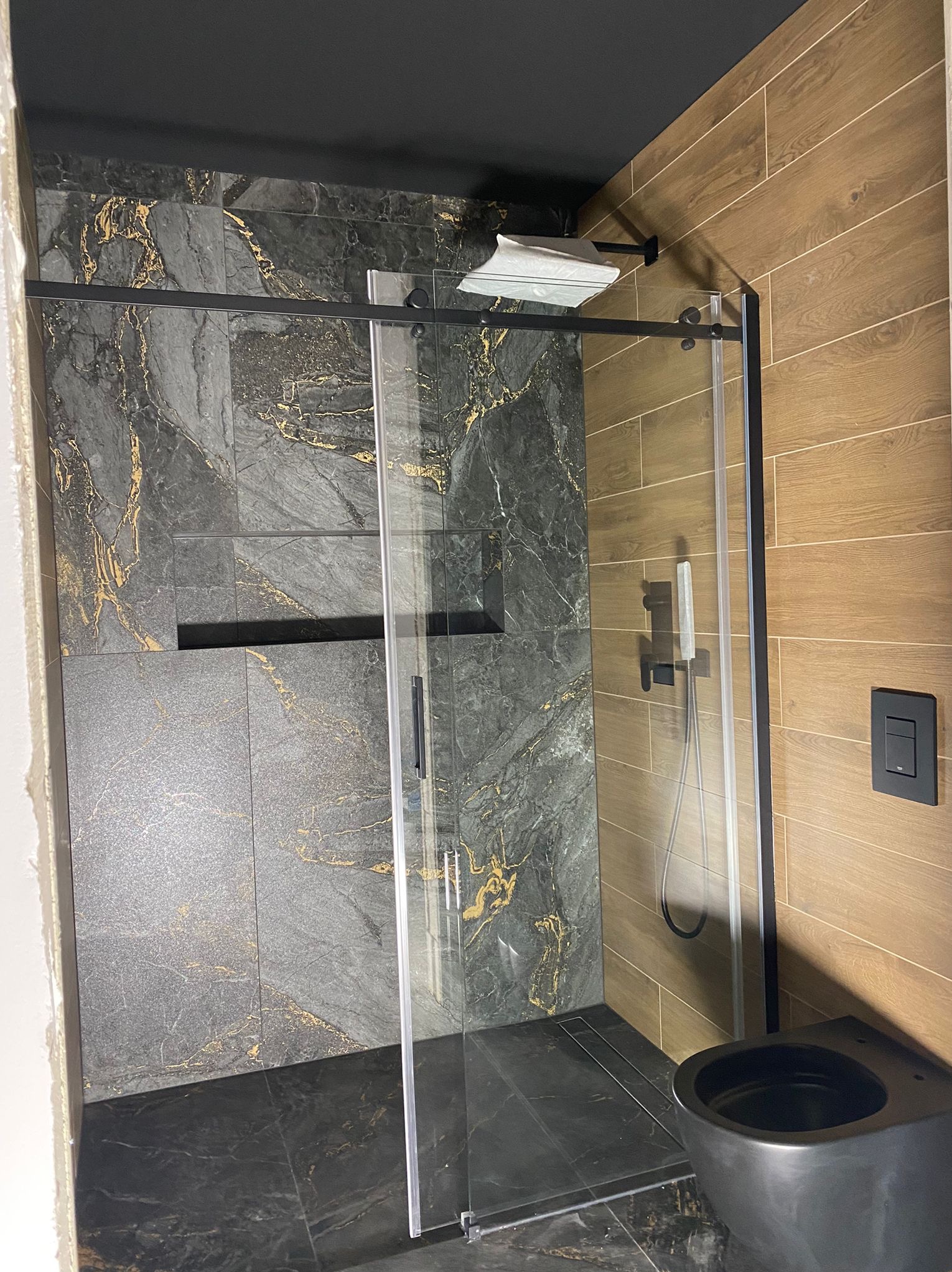 Nowoczesna łazienka po remoncie: kabina prysznicowa ze złotymi żyłkami na szarych płytkach, drewnopodobne płytki na ścianie, czarna armatura i sufit.
