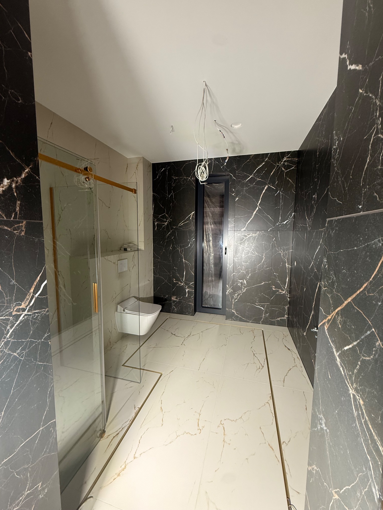 Elegancka łazienka z kabiną prysznicową ze złotymi detalami, marmurowe płytki ścienne w odcieniach czerni i bieli, biała podłoga z akcentami złota. Widoczne niedokończone prace.