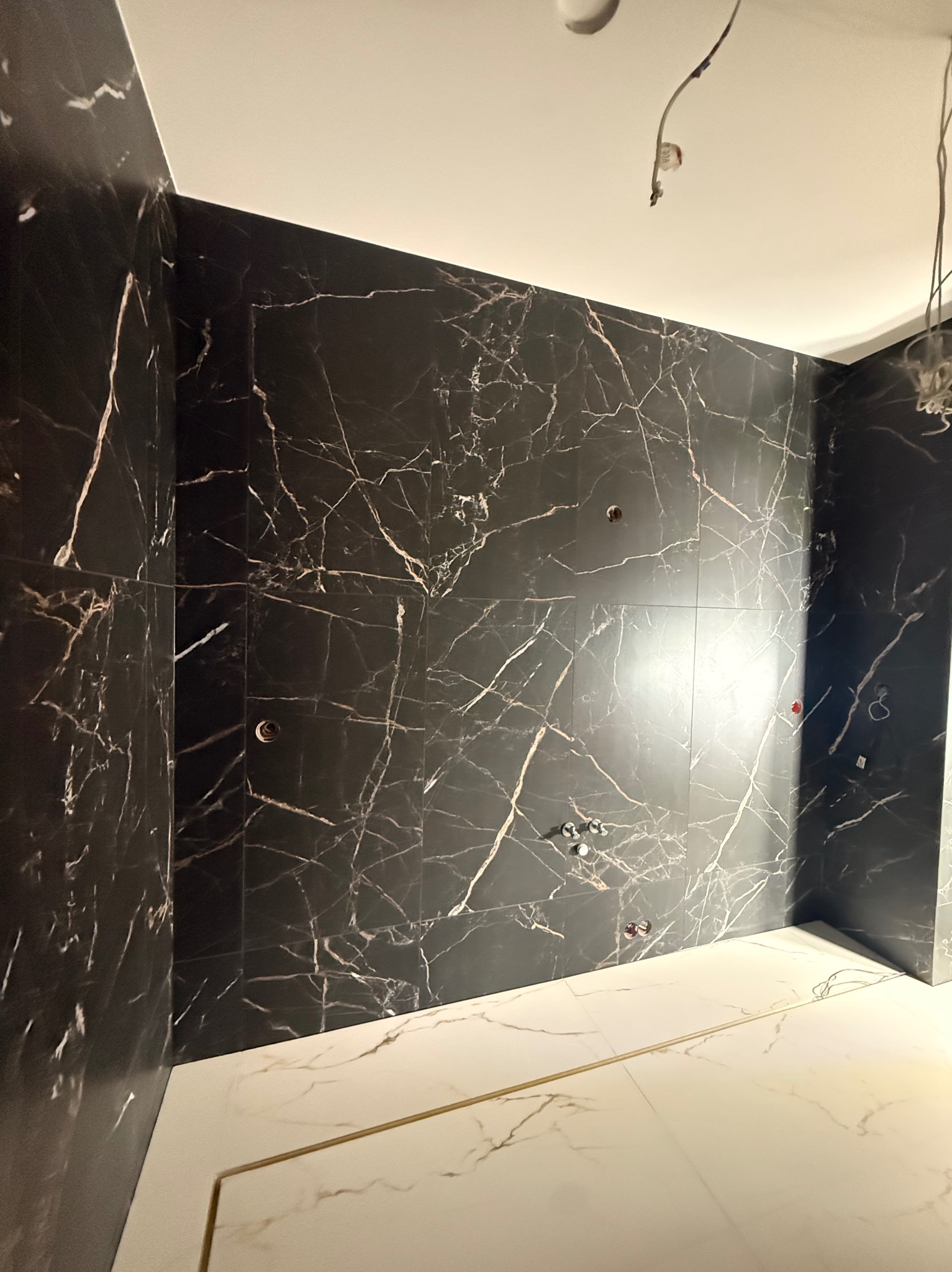 Wykończona łazienka z czarnymi płytkami imitującymi marmur na ścianach i białymi płytkami na podłodze. Widoczne instalacje wodne i elektryczne przed montażem armatury.